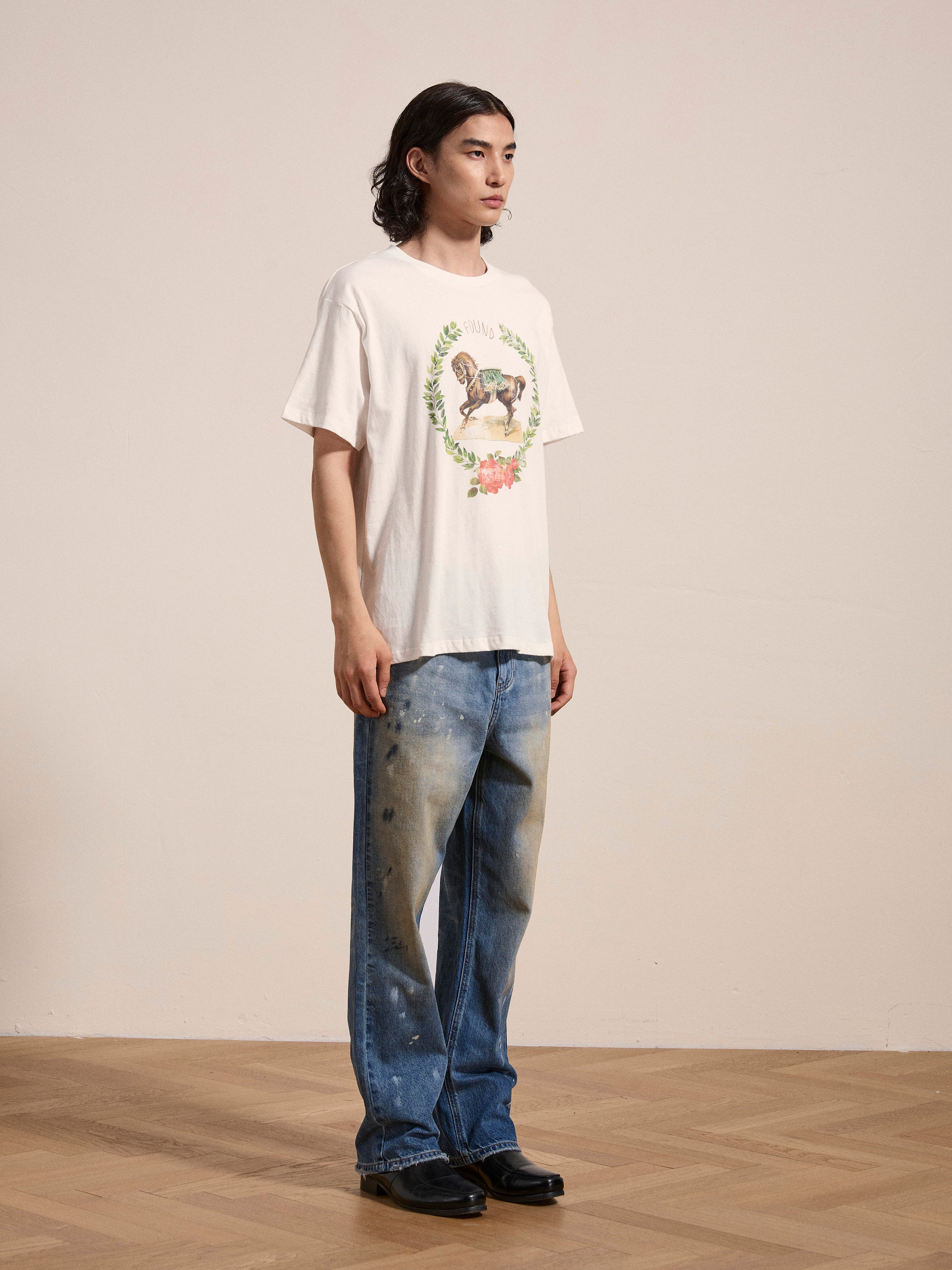 A person with long hair stands indoors on a wooden floor, wearing a white graphic T-shirt from FOUND's "Rodeo Tee" collection, baggy blue jeans with paint stains, and black shoes. The light-colored wall in the background enhances the vintage ambiance of the scene.