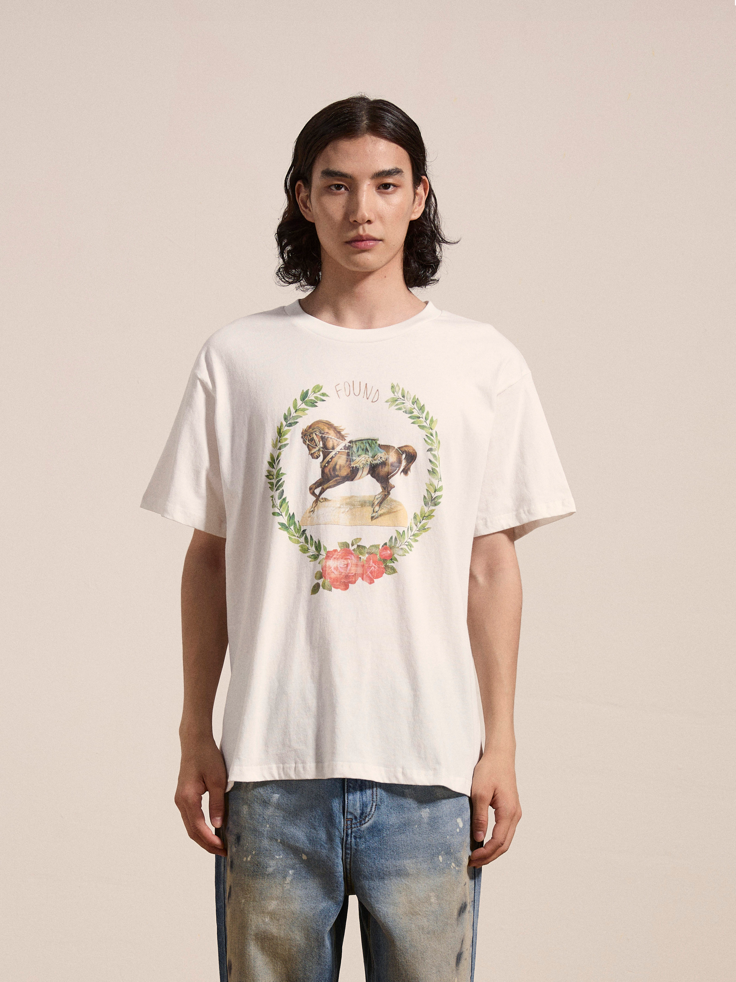 A person with long hair wears a white FOUND Rodeo Tee that offers a vintage vibe with floral wreath and horse graphics, paired with blue jeans, standing against a plain background.