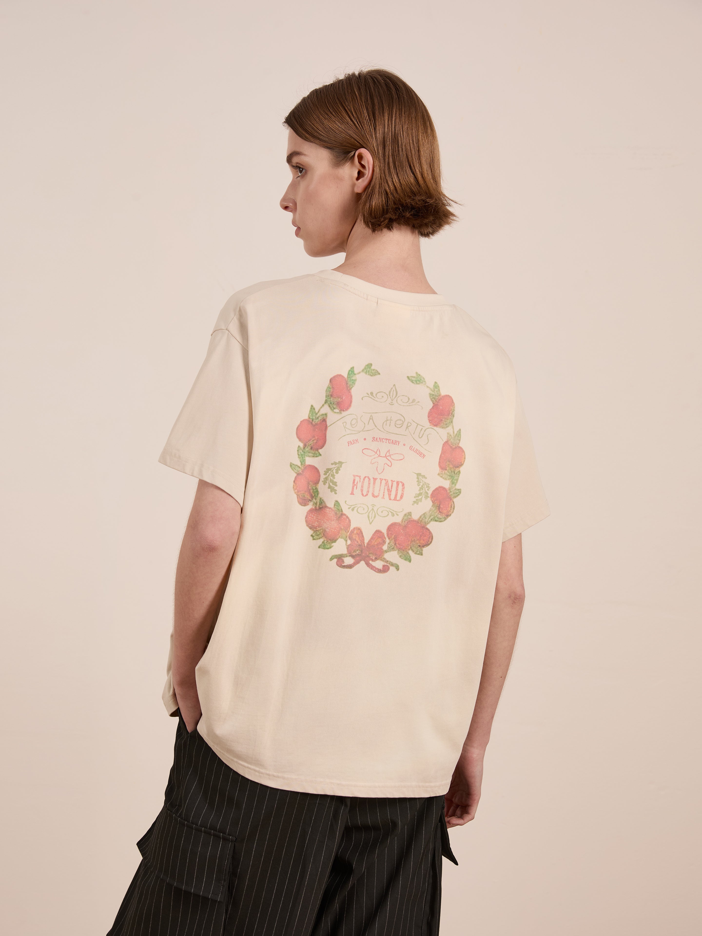 A short-haired person stands sideways, dressed in a Rose Garden Tee by FOUND, featuring a floral wreath graphic on the back, and black pinstripe pants.