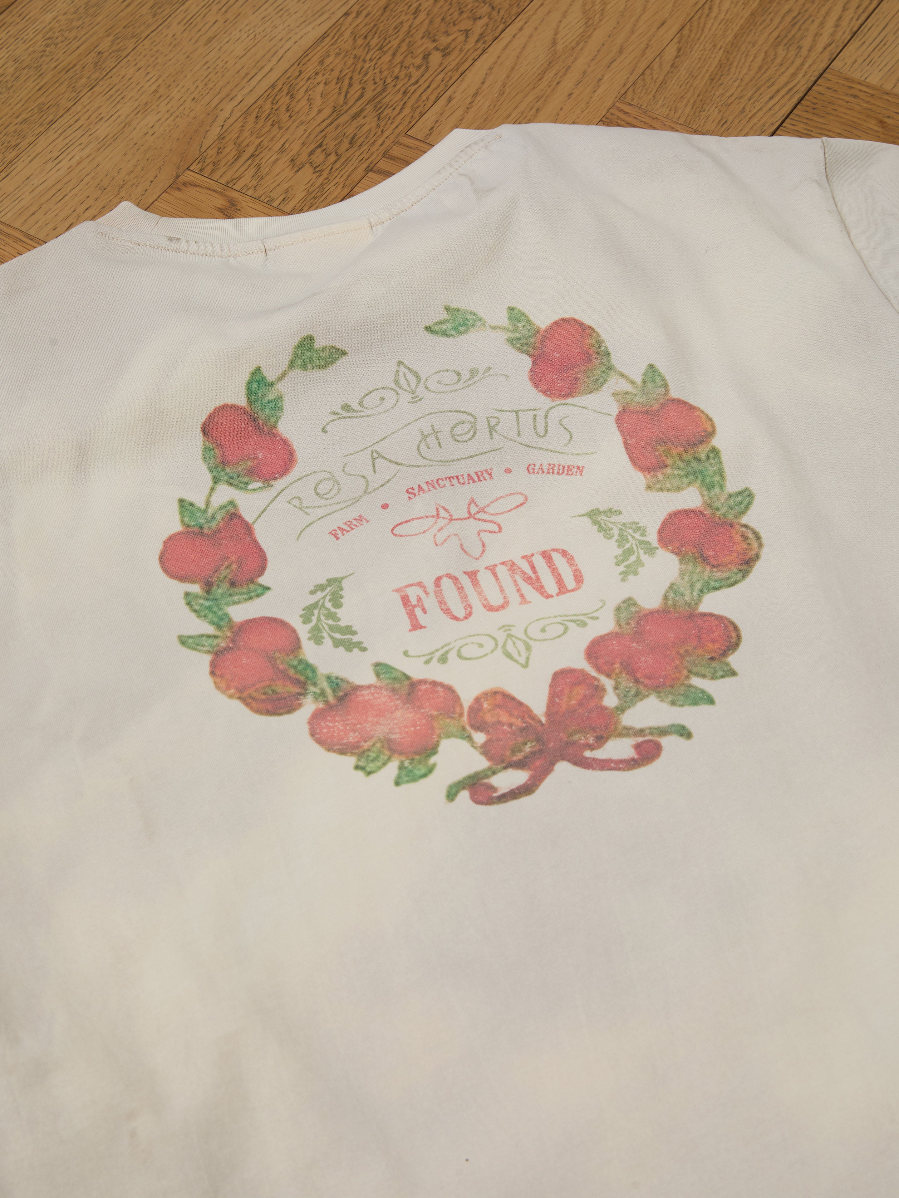 The Rose Garden Tee from FOUND features a floral wreath graphic with "Rosa Hortus," "Find Sanctuary + Garden," and "Found" on a white t-shirt that's laid out on a wooden floor, exuding vintage charm perfect for timeless elegance seekers.