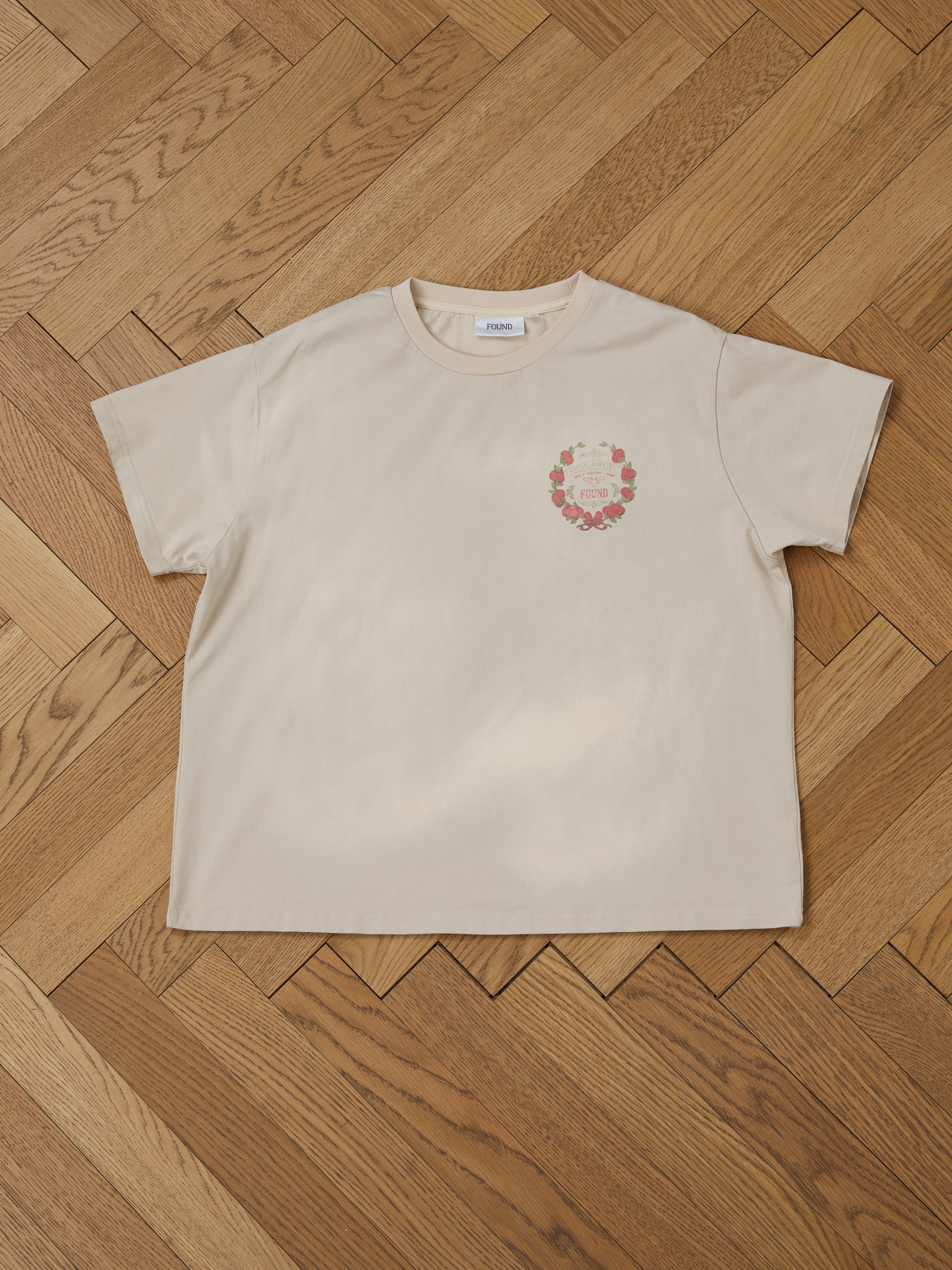 The Rose Garden Tee by FOUND, with its beige hue and floral wreath graphic paired with text on the left chest, exudes vintage charm while resting on a wooden herringbone-patterned floor.