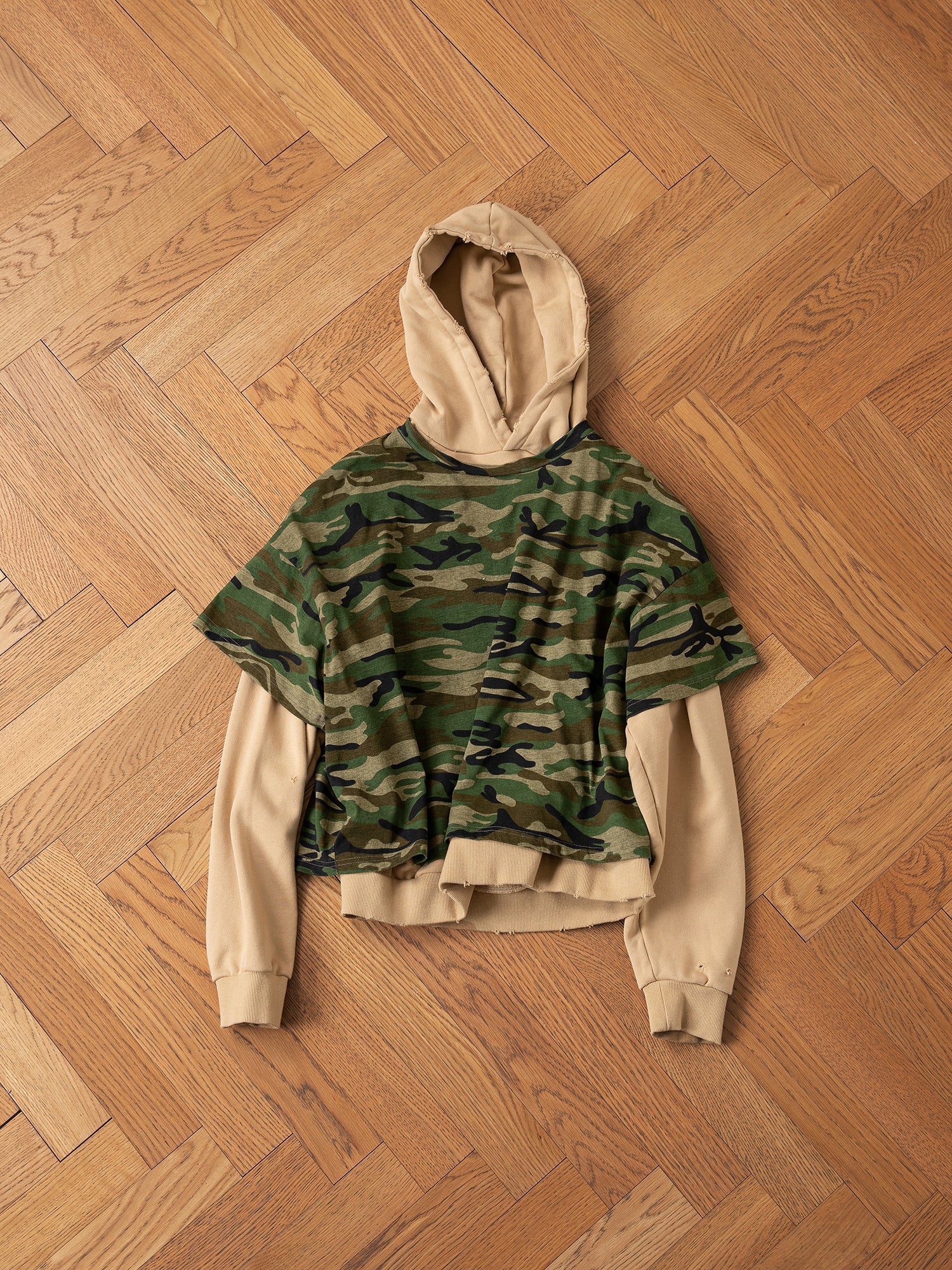 A Double Layer Hoodie from FOUND, made with French terry cotton, is elegantly combined with a green camouflage short-sleeved shirt to achieve a vintage style, all set against a wooden parquet floor.