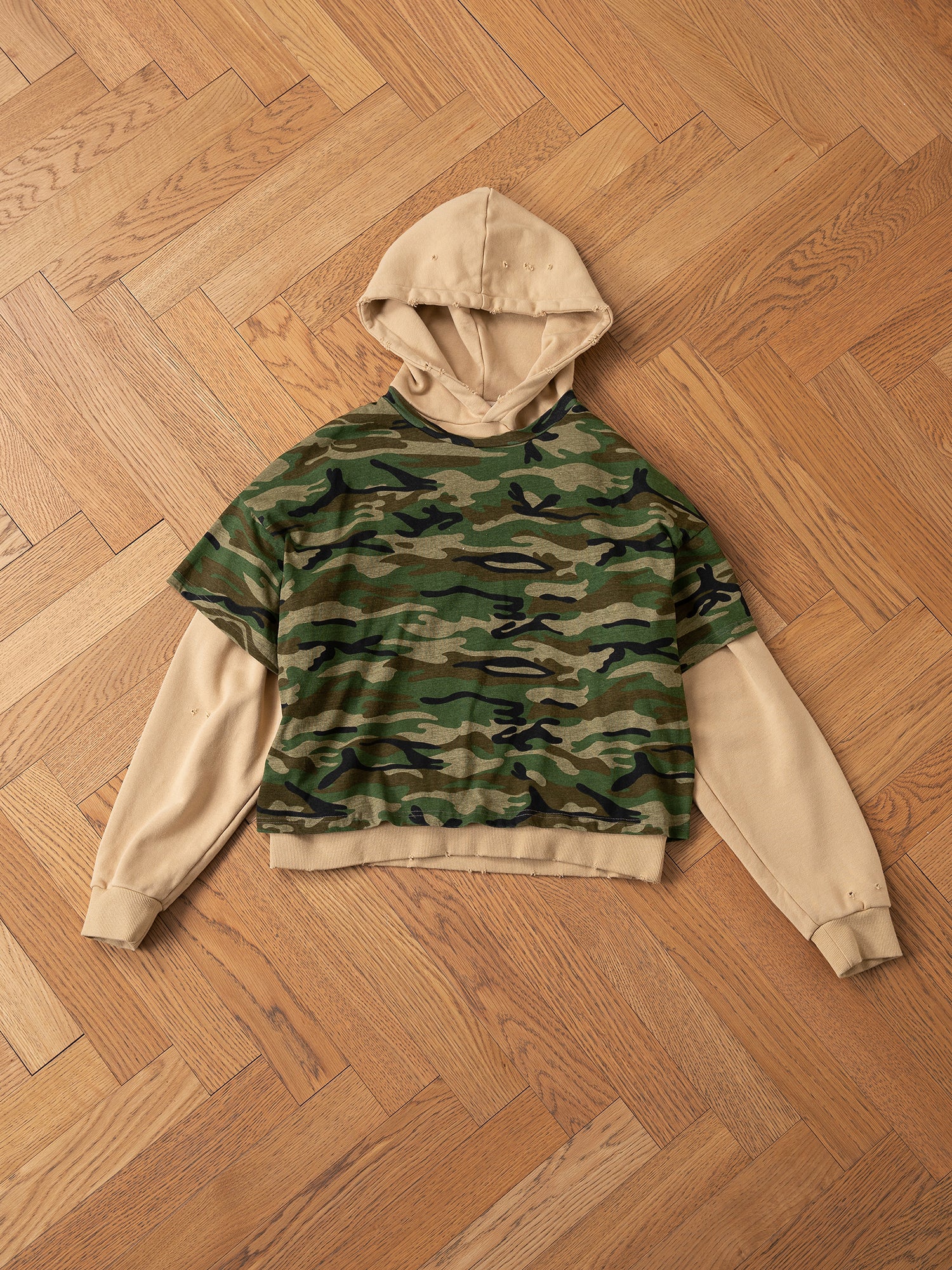 A Double Layer Hoodie by FOUND, showcasing a vintage camouflage pattern with beige sleeves and hood, crafted from soft French terry cotton, is arranged flat on a wooden parquet floor.