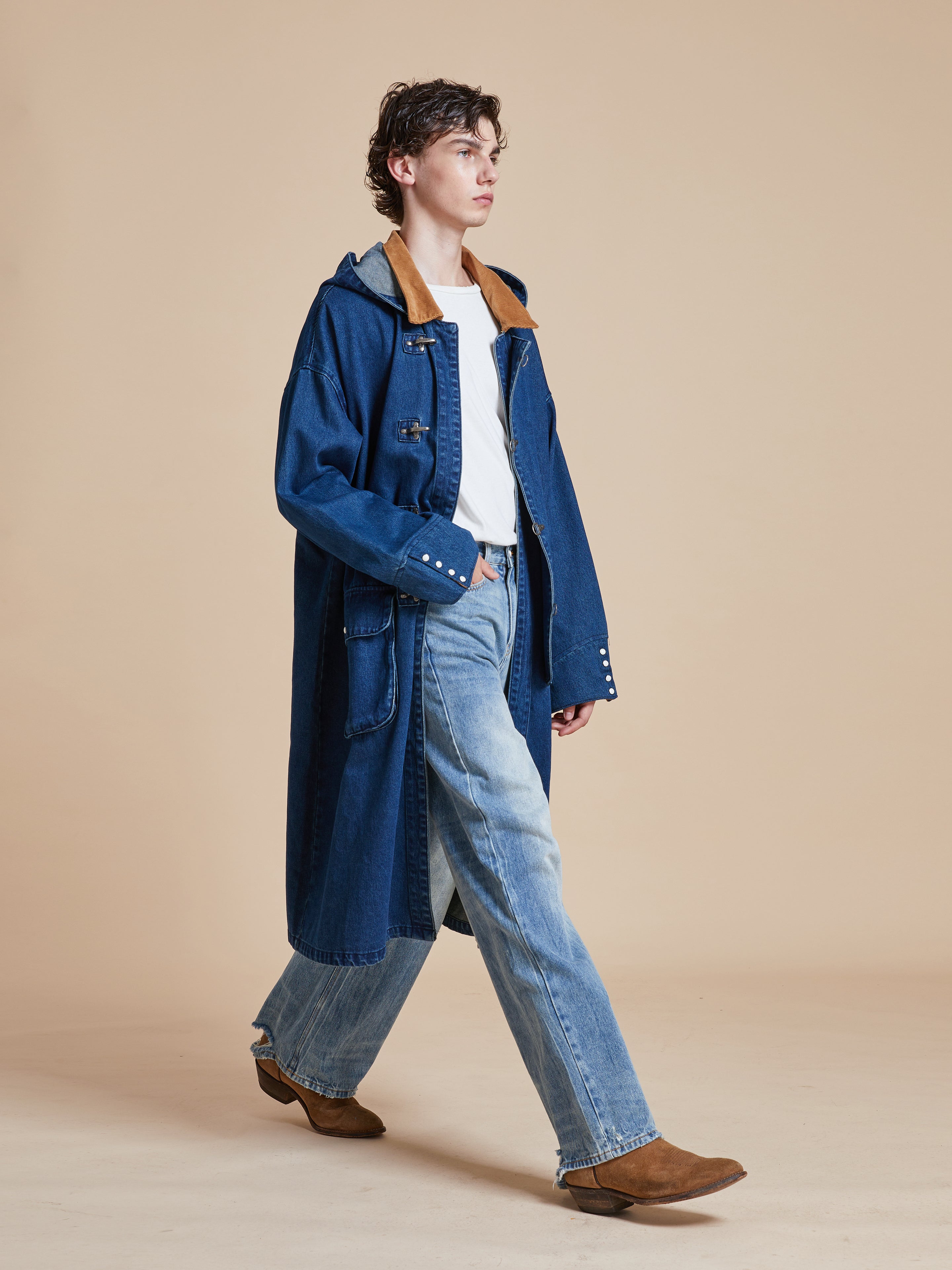 A man in a Sargasso Denim Buckle Coat and jeans walking on a beige background.