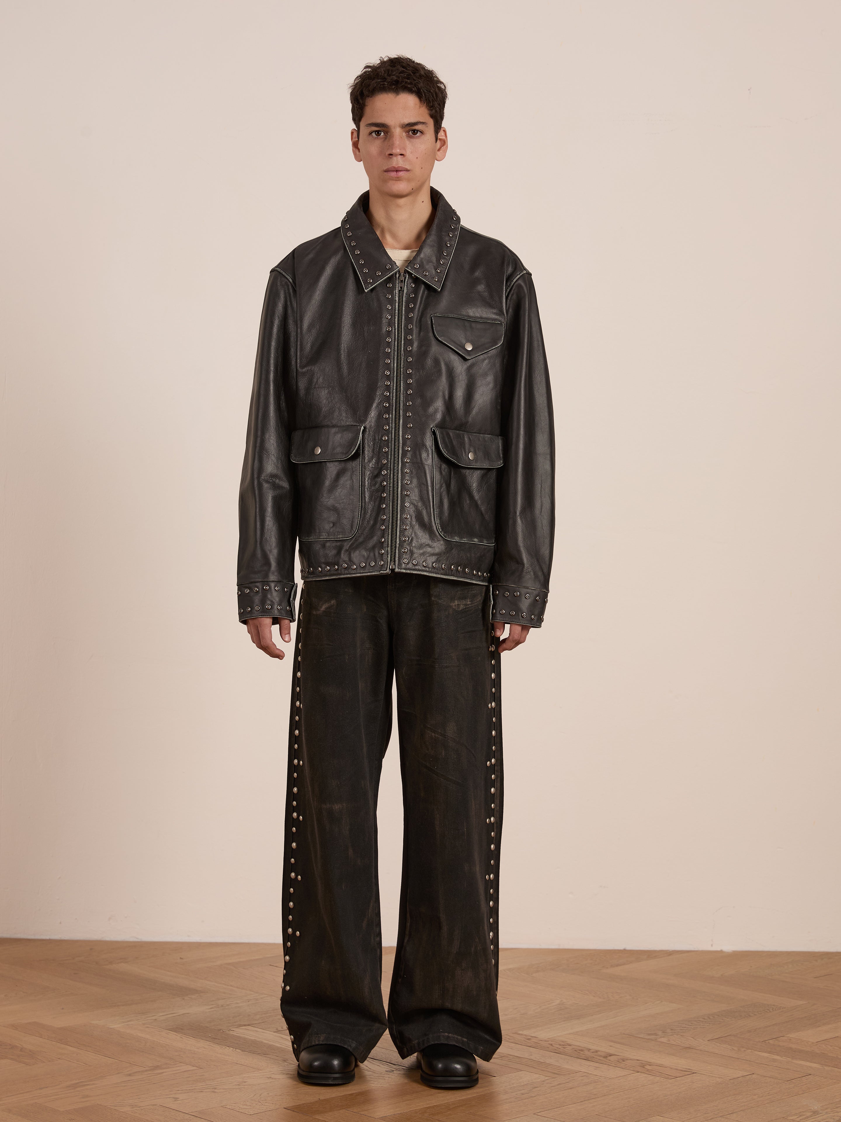 A person stands on a wooden floor against a plain wall, wearing the FOUND Studded Leather Pocket Jacket and wide-leg pants adorned with silver studs.