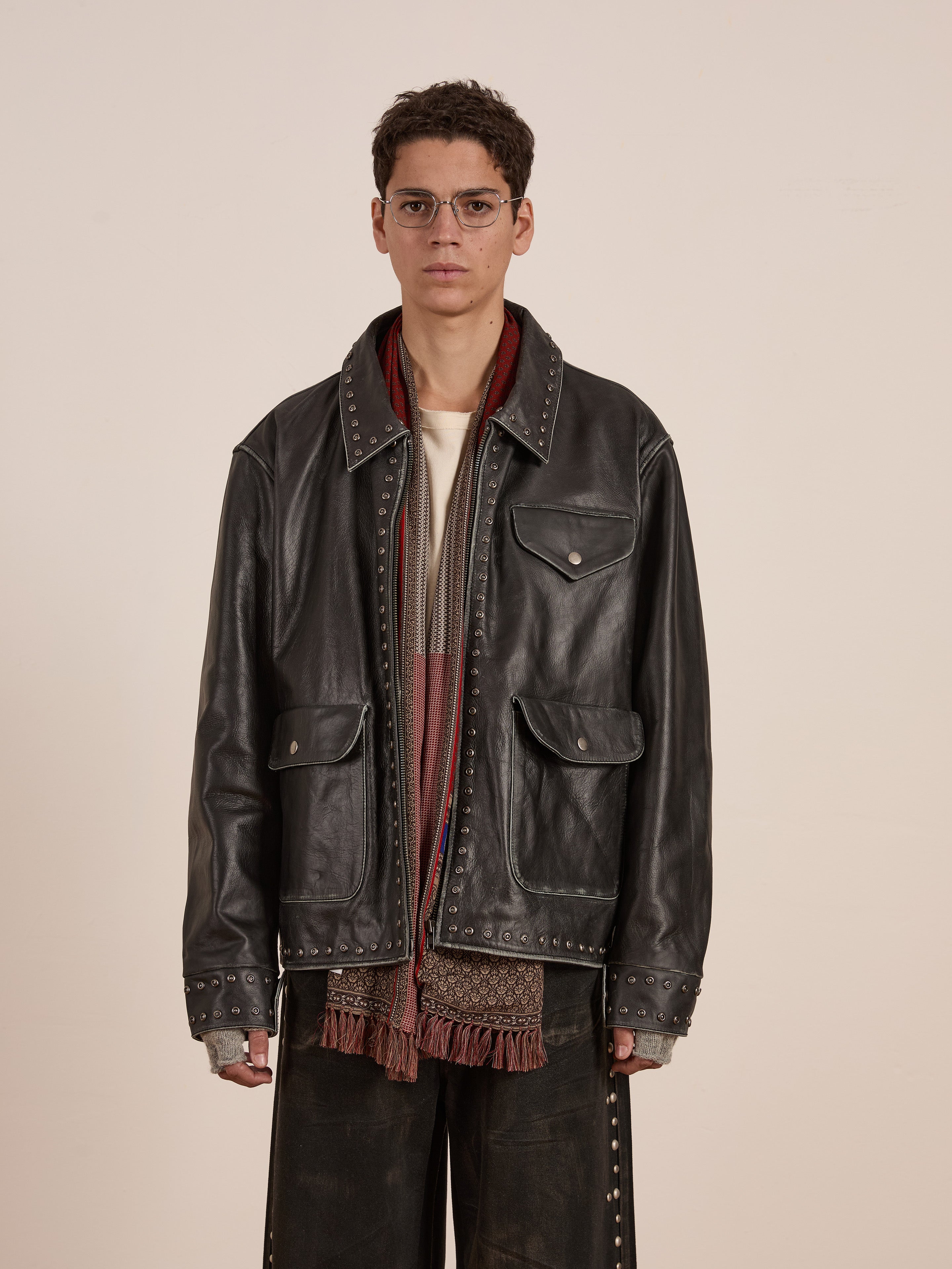 Wearing FOUND's Studded Leather Pocket Jacket, a person stylishly pairs it with glasses, a red fringed scarf, and dark pants while standing confidently against a plain background.