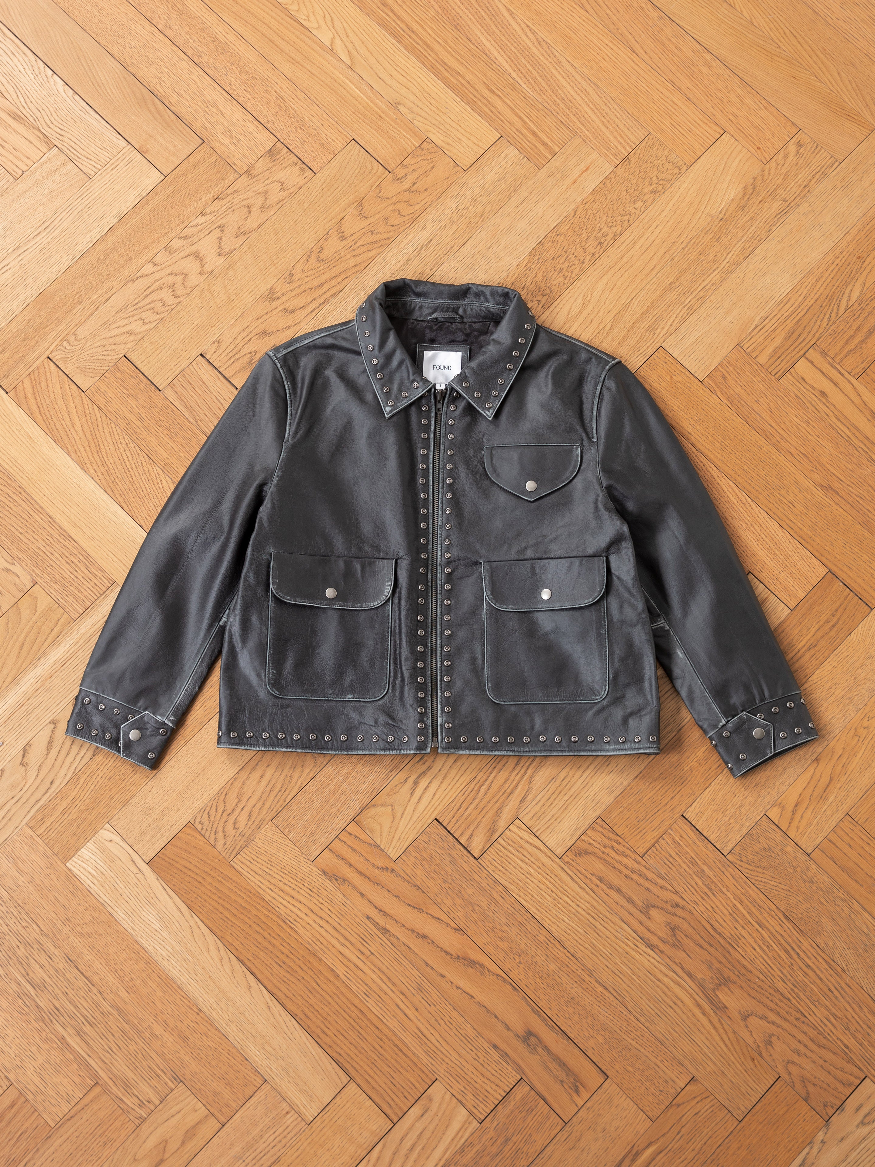 The Studded Leather Pocket Jacket by FOUND is stylishly showcased on a wooden herringbone-patterned floor.