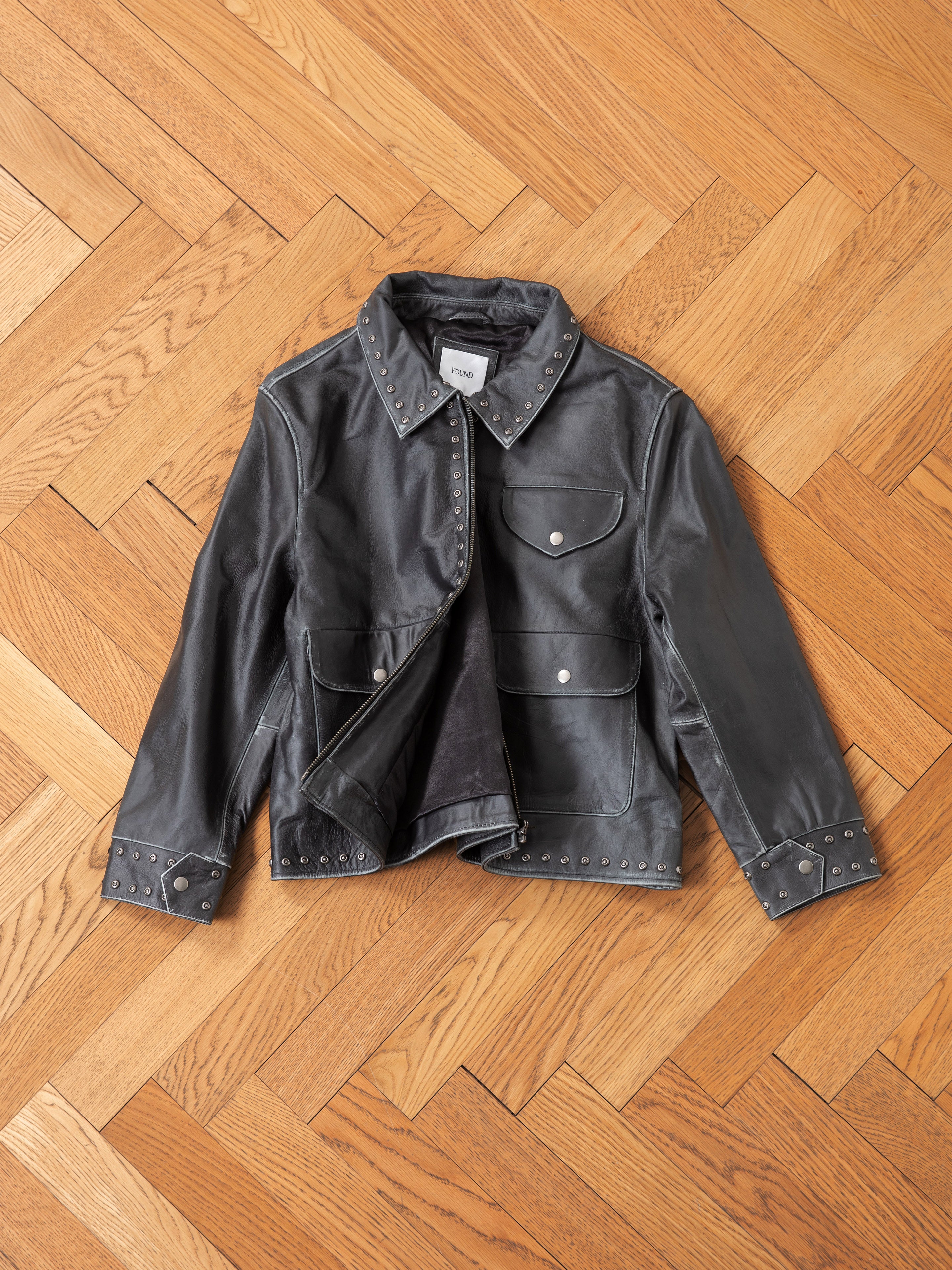 The Studded Leather Pocket Jacket by FOUND, a vintage-style black leather piece with buttoned pockets and stud detailing, ideal for any unisex wardrobe, is laid flat on a wooden herringbone floor.