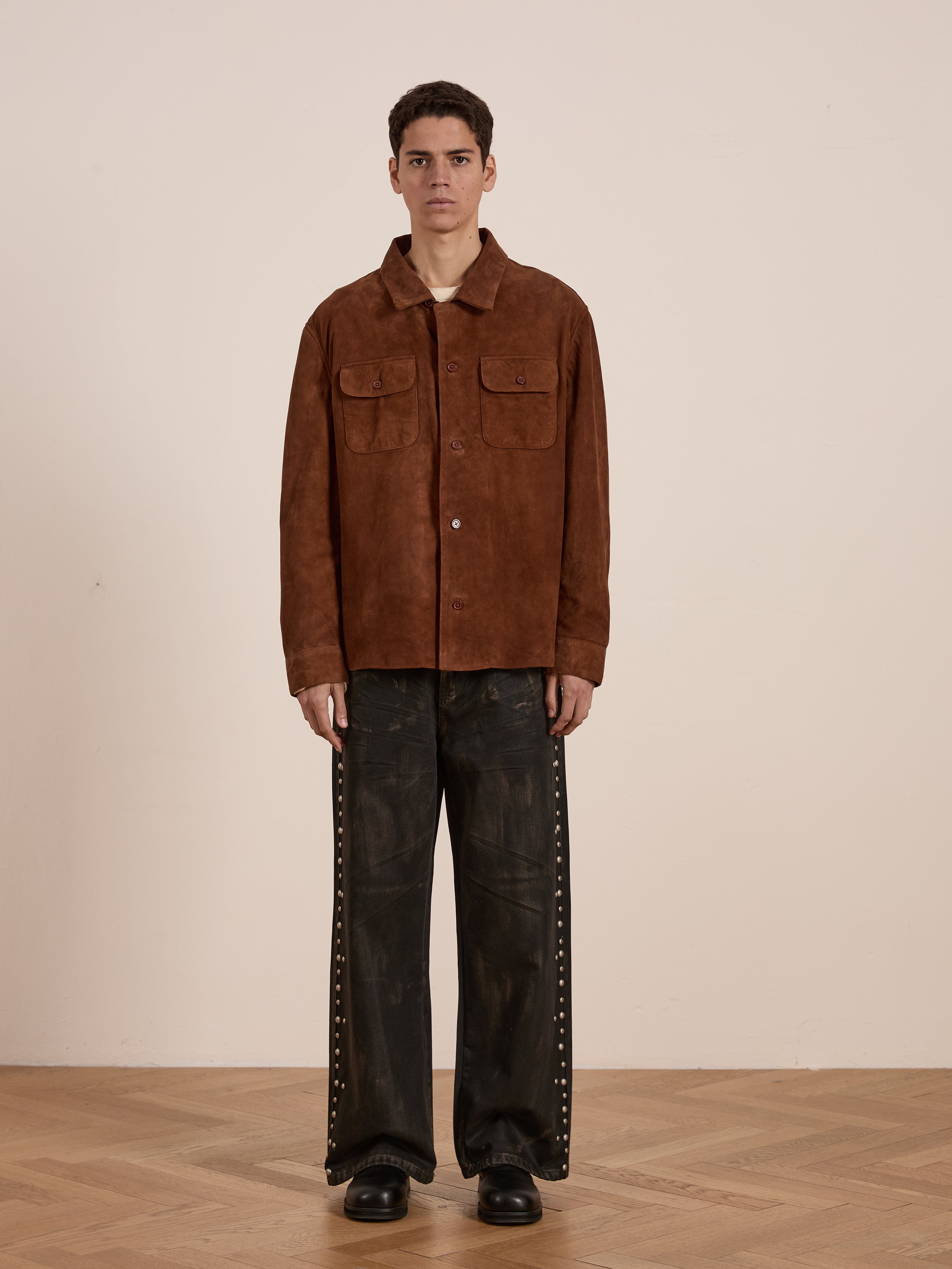 A person stands indoors in the Suede Camp Shirt by FOUND, a relaxed genuine goat suede leather jacket. They pair it with wide-legged dark pants featuring rivet details and black shoes, all set against a plain background with a wooden floor.