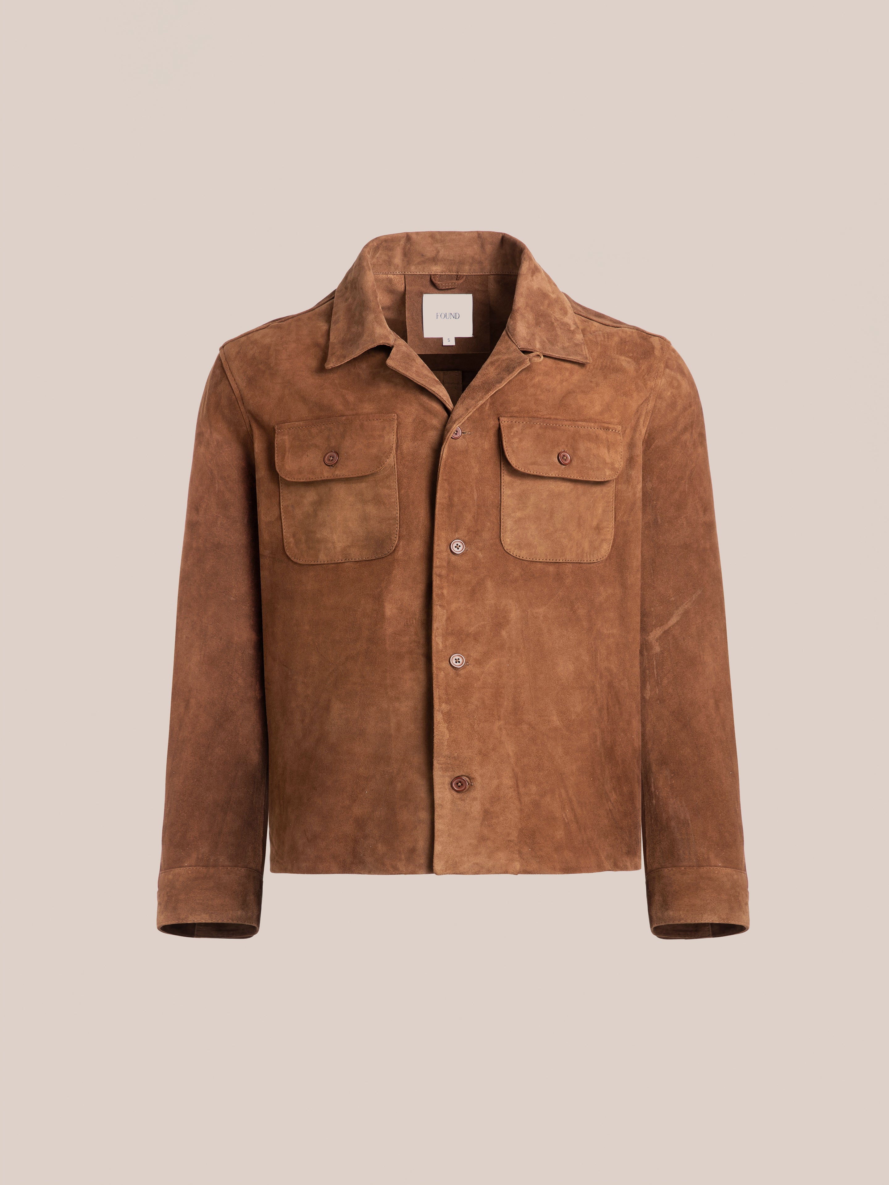 The FOUND Suede Camp Shirt features a button-up front, relaxed silhouette, two chest pockets, and a folded collar. Made from genuine goat suede leather in brown, it is set against a plain beige background.