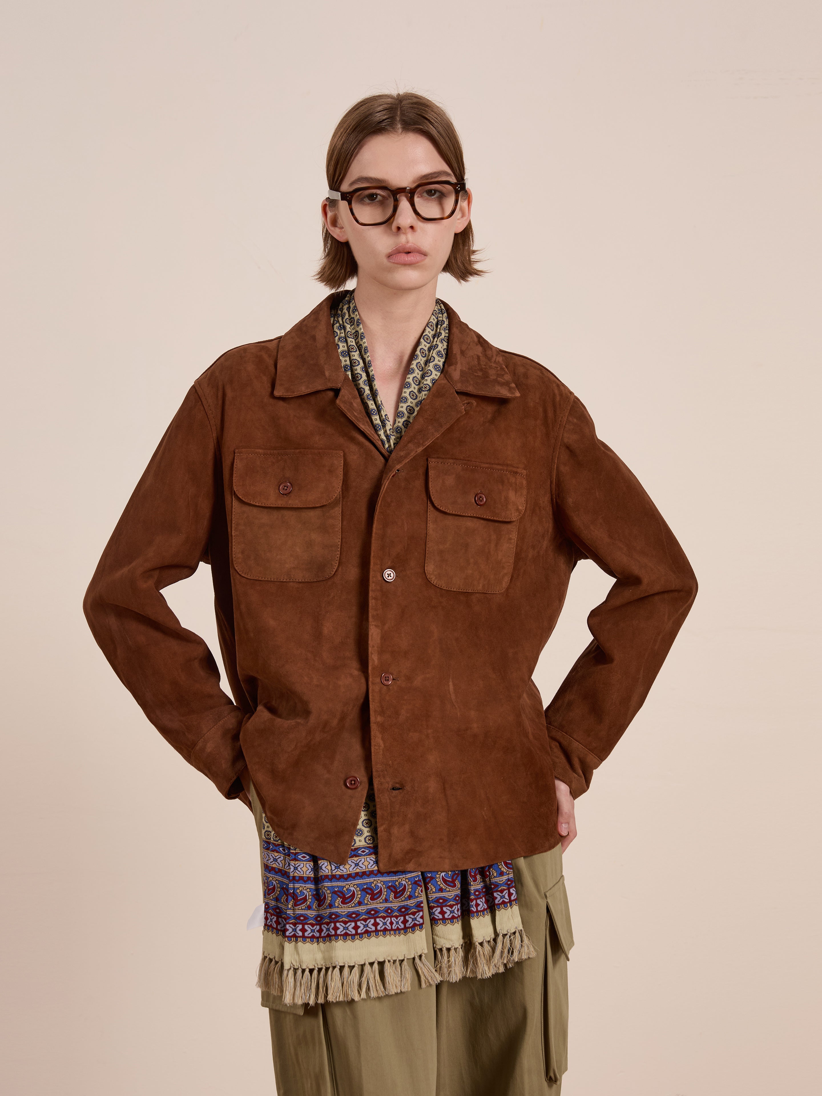 A person with long hair and glasses stands against a plain backdrop, wearing FOUND's brown Suede Camp Shirt styled like workwear, complete with front pockets. An elegantly draped patterned scarf accents the look, paired with olive pants.