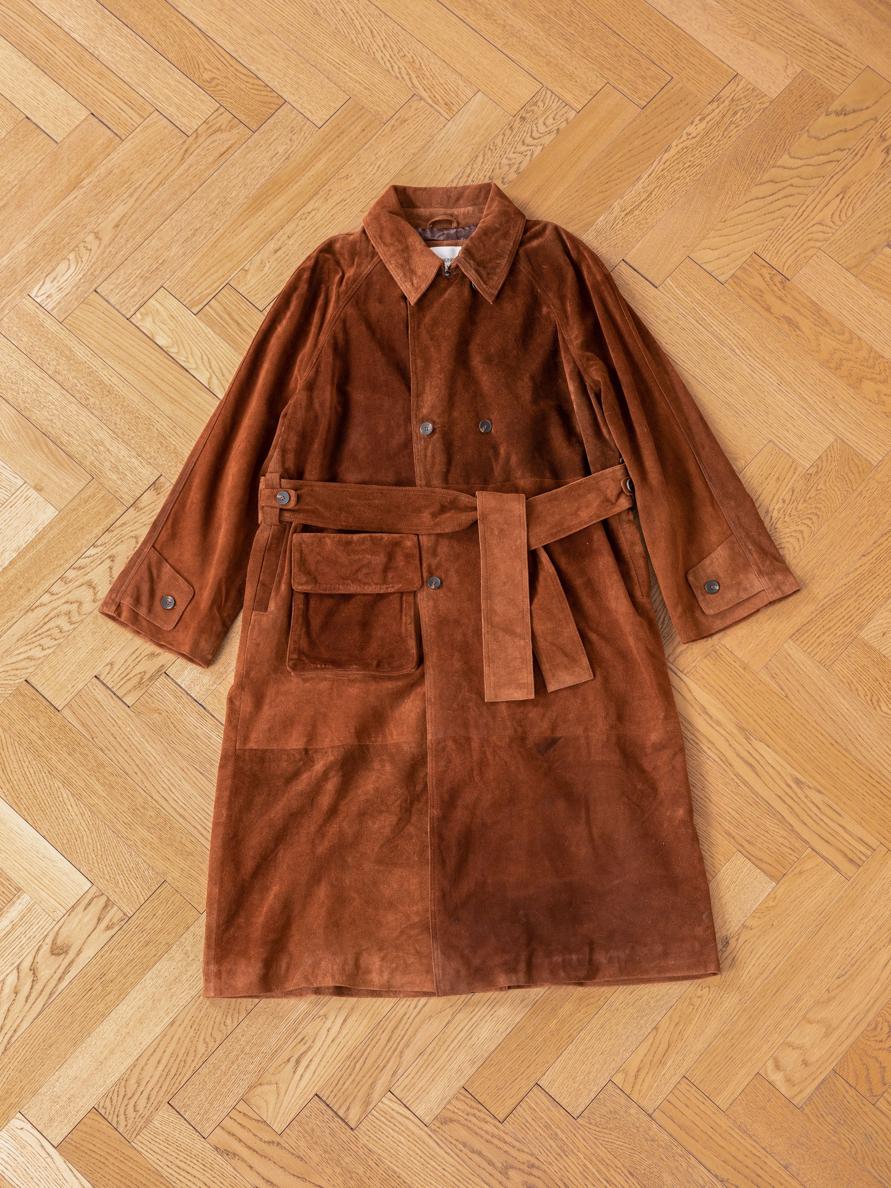 The Suede Leather Trench Coat by FOUND, made from brown suede leather, features a belt and brown tortoise buttons. It is ethically crafted with a front pocket for convenience and displayed on a wooden parquet floor.