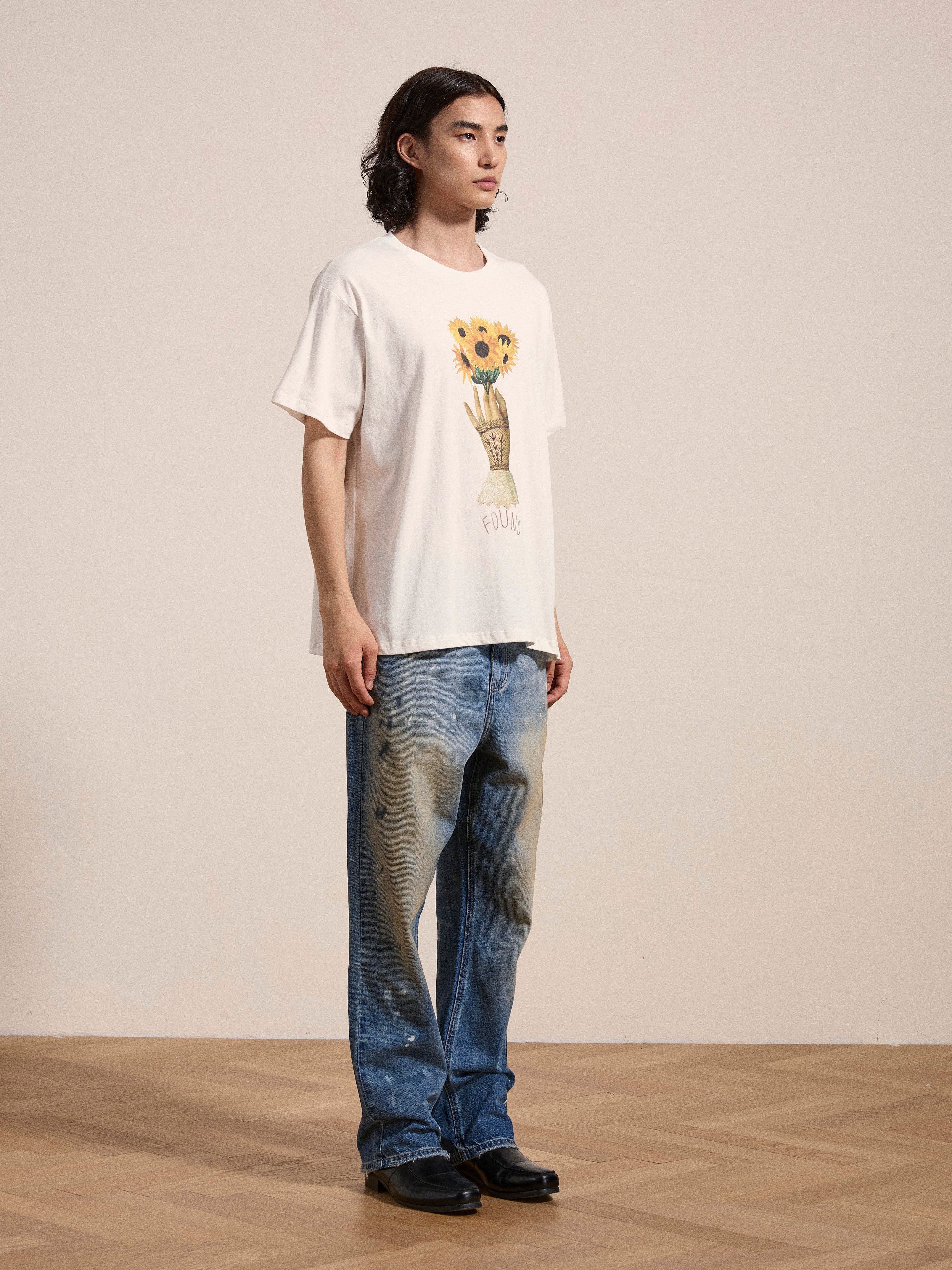 In a neutral-toned room, a person stands wearing the FOUND Sunflower Tee adorned with a floral graphic and blue jeans featuring a washed-out effect, giving off a vintage feel. Their shoulder-length hair complements their choice of black shoes.