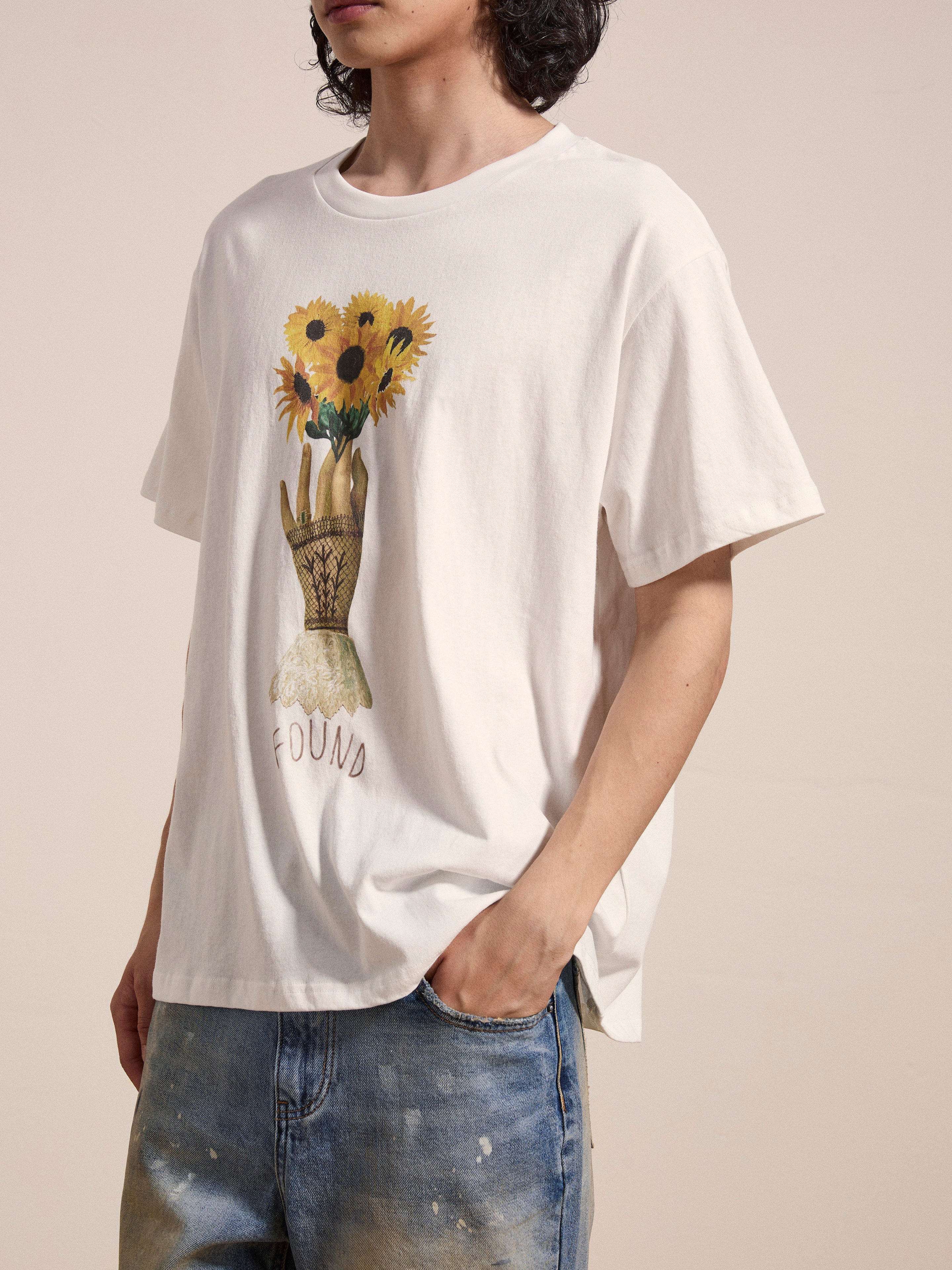 A person is wearing the Sunflower Tee by FOUND, a vintage feel white t-shirt featuring a floral graphic of a hand holding sunflowers with "FOUND" printed below it. They are also wearing blue jeans.