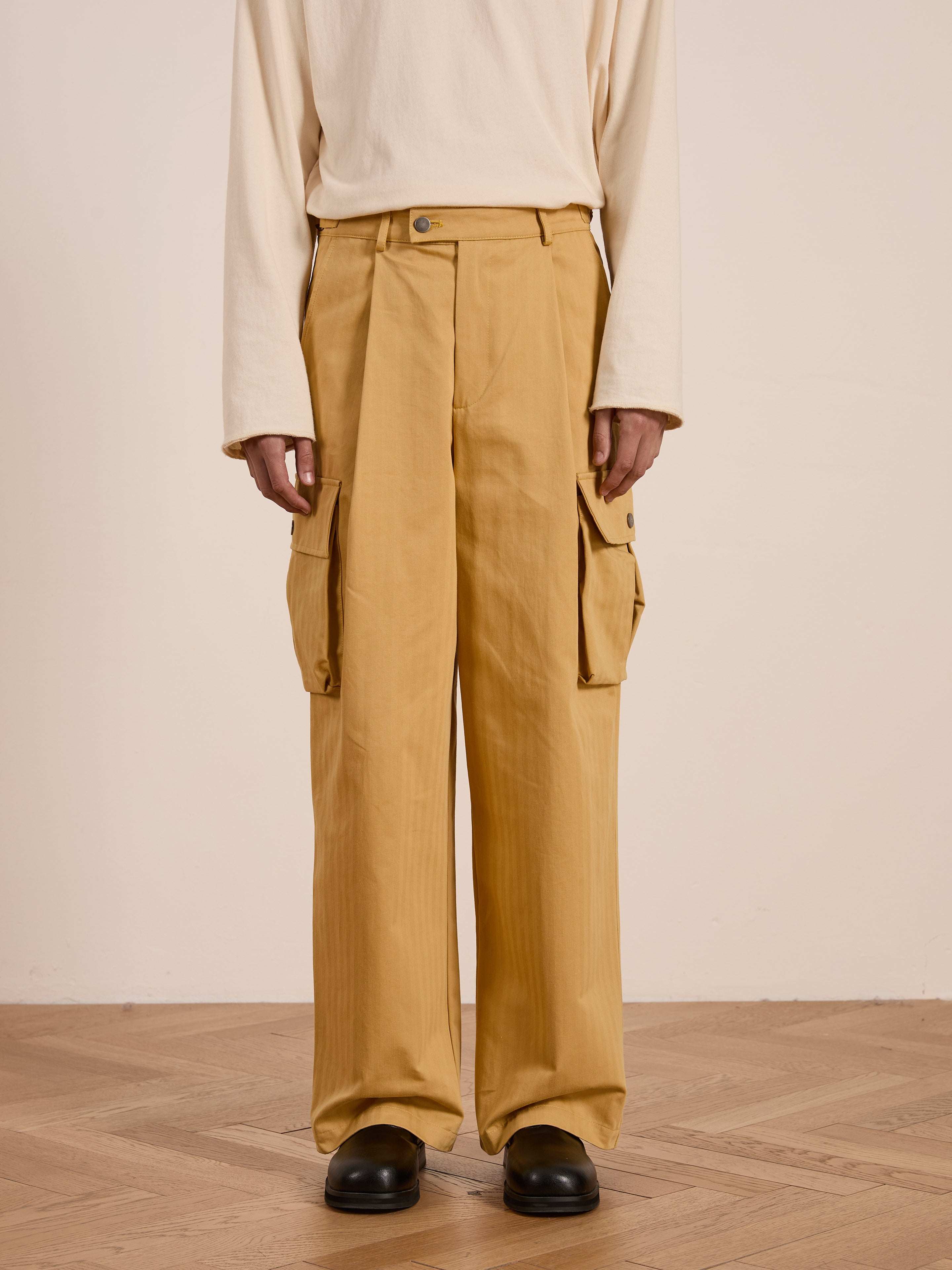 A person wearing a beige long-sleeve shirt and the FOUND Sunlight Twill Cargo Pants, which are unisex mustard cargo pants with a relaxed fit and large pockets, stands on a wooden floor.