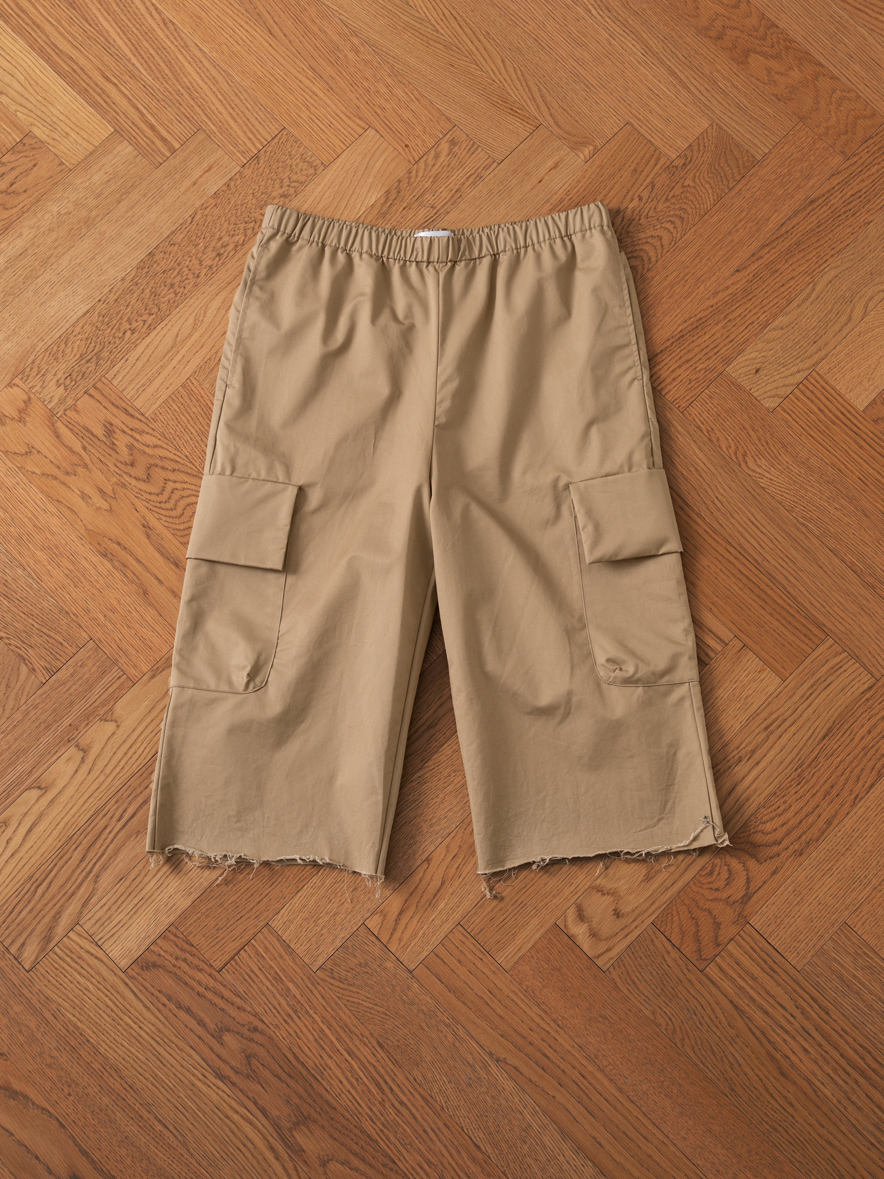 A pair of Long Twill Cargo Shorts by FOUND in beige, featuring frayed hems and made from lightweight cotton fabric, laid flat on a wooden herringbone floor, showcasing the dual cargo pockets.