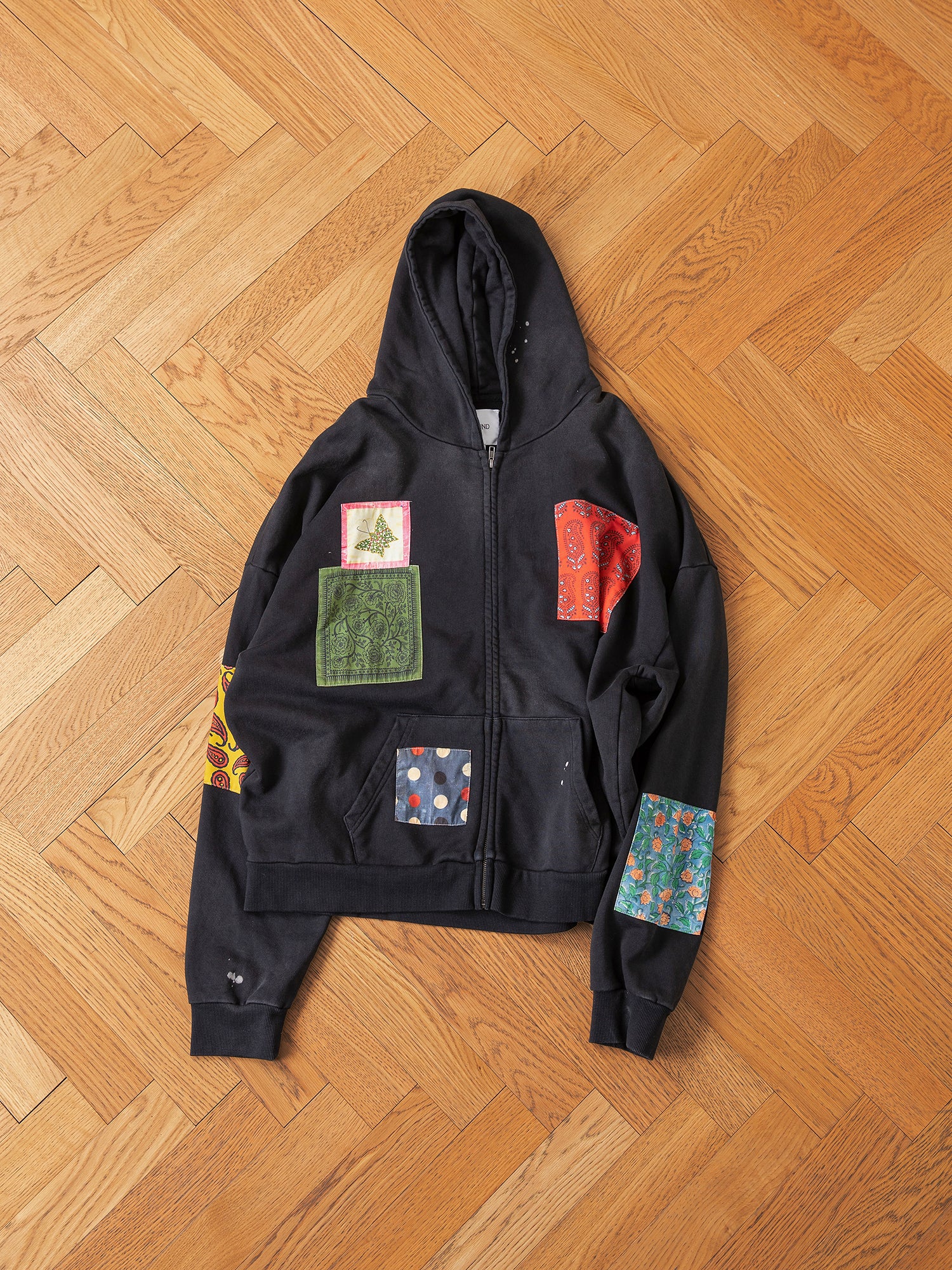 Displayed on a wooden herringbone floor is the Tapestry Patchwork Zip Up Hoodie by FOUND, a vintage-inspired black hoodie featuring an oversized fit with an array of colorful patches.