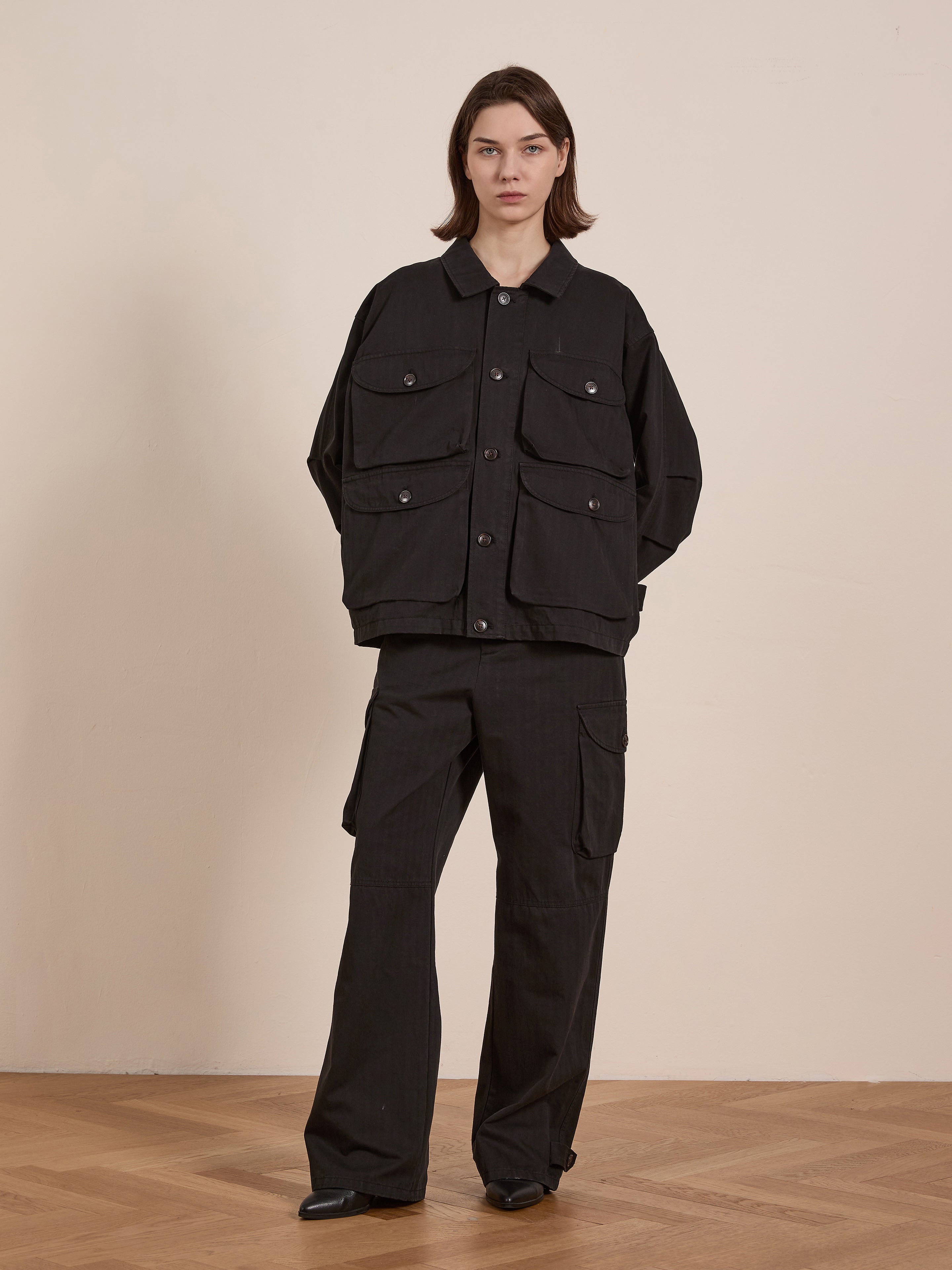 A person stands on a wooden floor wearing a black button-up jacket with large pockets, exuding a vintage look. Paired with FOUND's Utility Herringbone Cargo Pants, the ensemble showcases durable cotton twill fabric that combines fashion and function effortlessly.