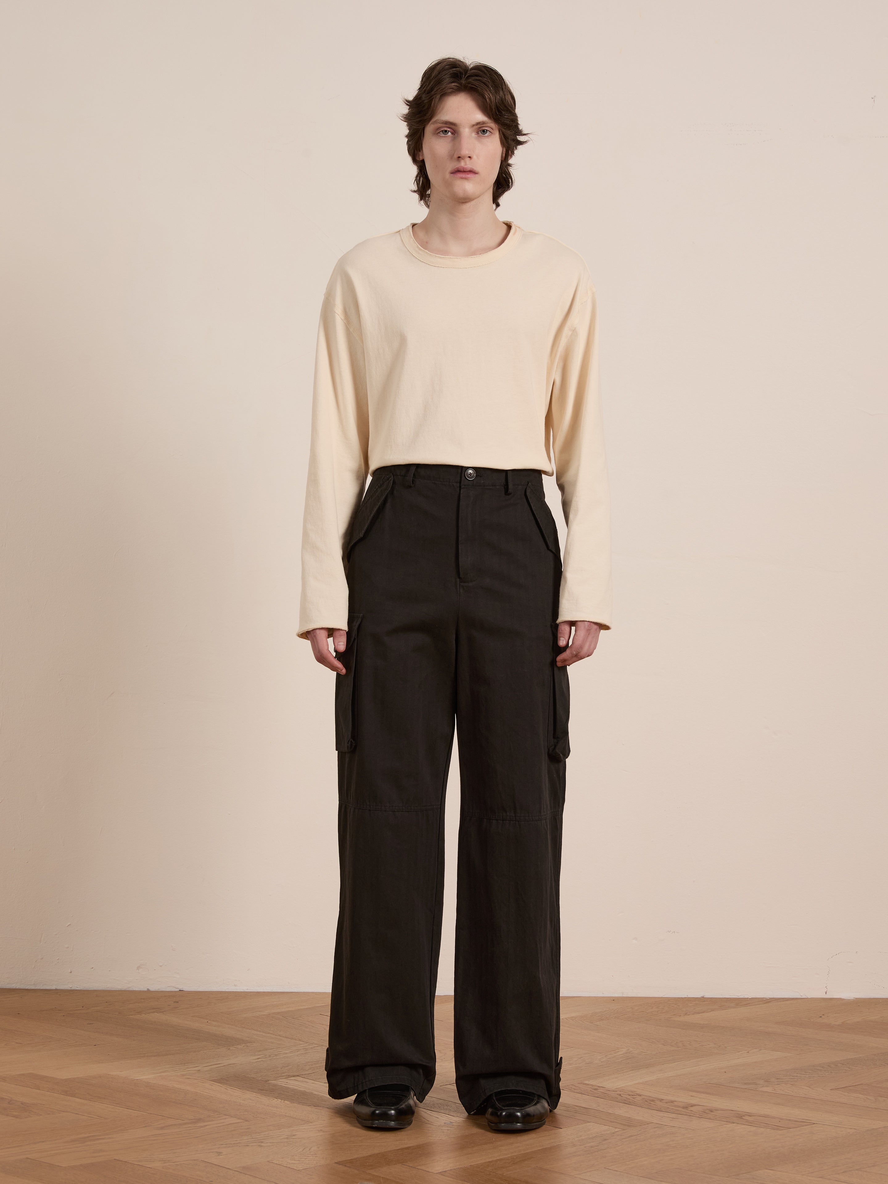 Indoors, someone wears a long-sleeve beige shirt with FOUND's black Utility Herringbone Cargo Pants, epitomizing a vintage look. The ensemble is completed with black shoes on a wooden floor.