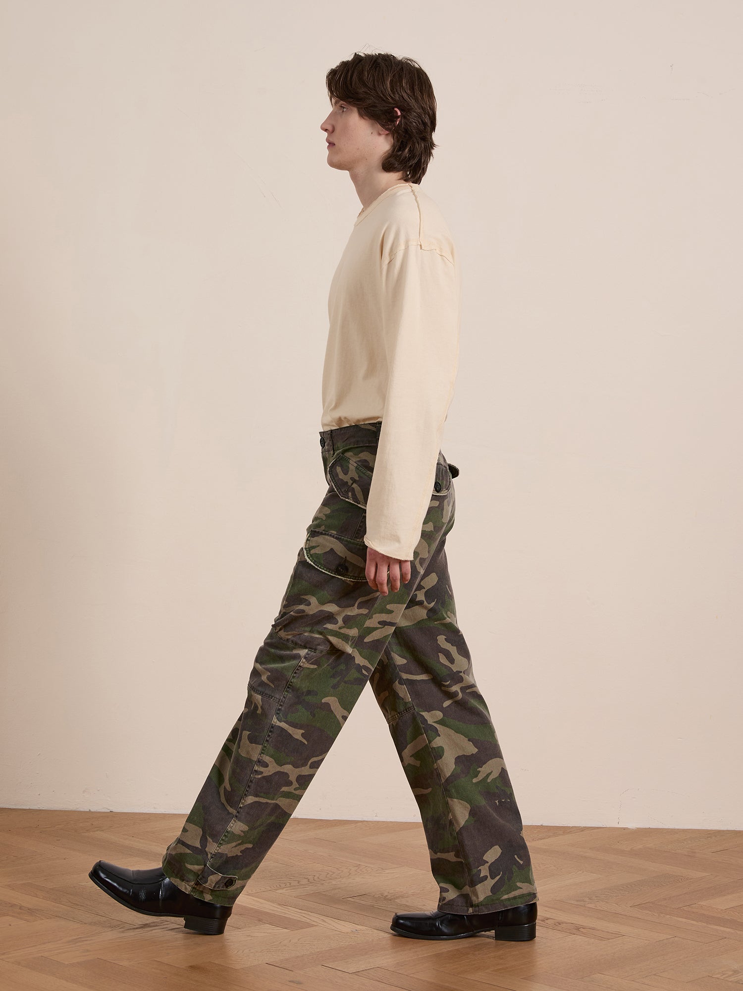 Against a plain wall, a person walks in profile wearing a cream long-sleeve shirt, Camouflage Twill Utility Cargo Pants by FOUND, and black shoes.