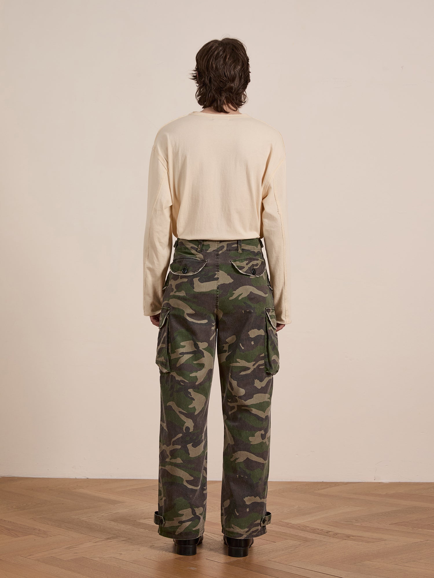 A person wearing a beige long-sleeve shirt and FOUND's Camouflage Twill Utility Cargo Pants stands on a wooden floor, facing away, exuding a vintage style with the stylish camo pattern.