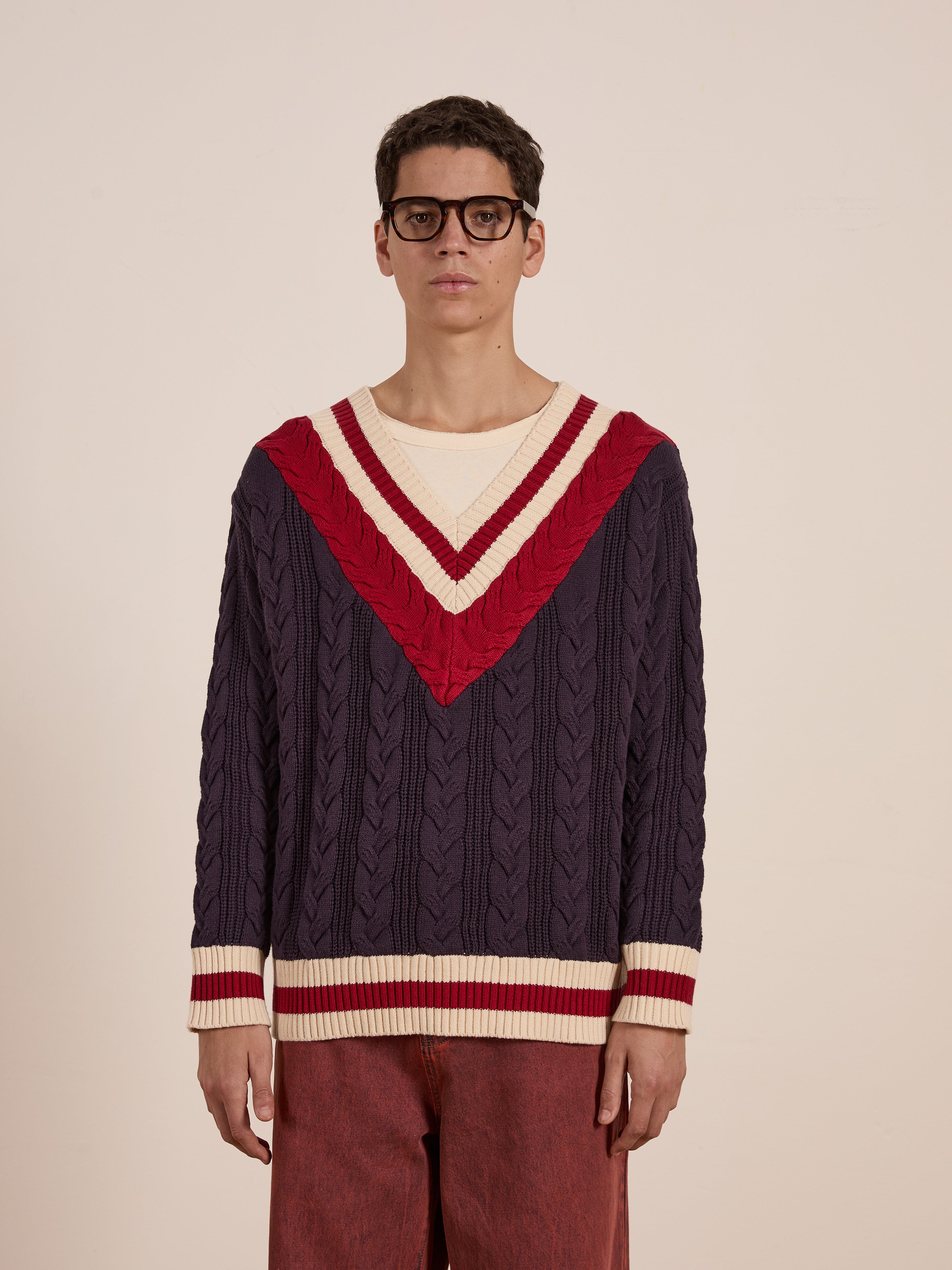 A person wearing glasses stands against a plain background, highlighting the navy and red FOUND V-Neck Cricket Sweater with a classic cable knit design, paired with reddish-brown pants.