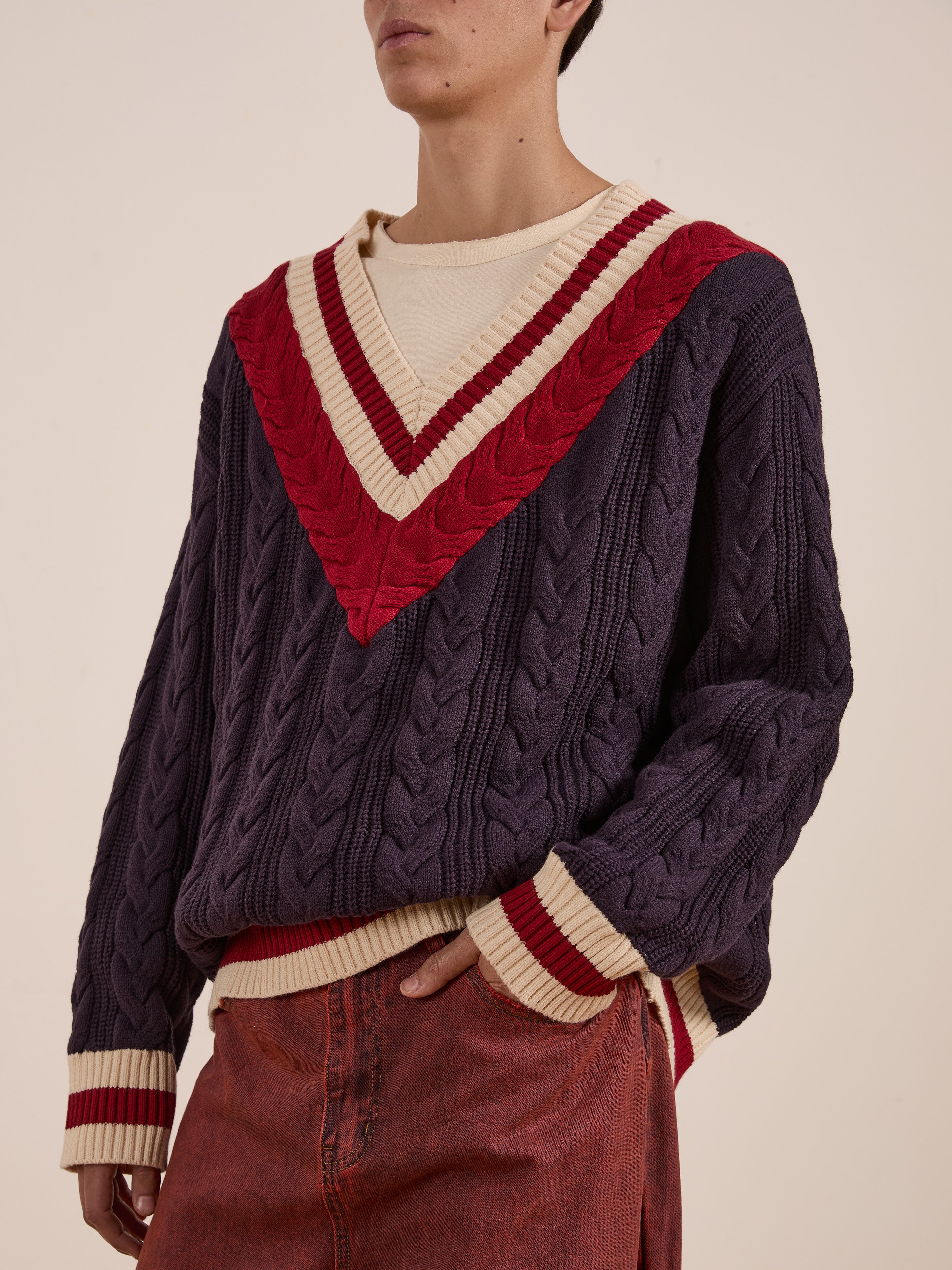 A person wears a stylish V-Neck Cricket Sweater by FOUND, featuring a cable-knit design with red and beige details, over a light cream shirt and vibrant red trousers.