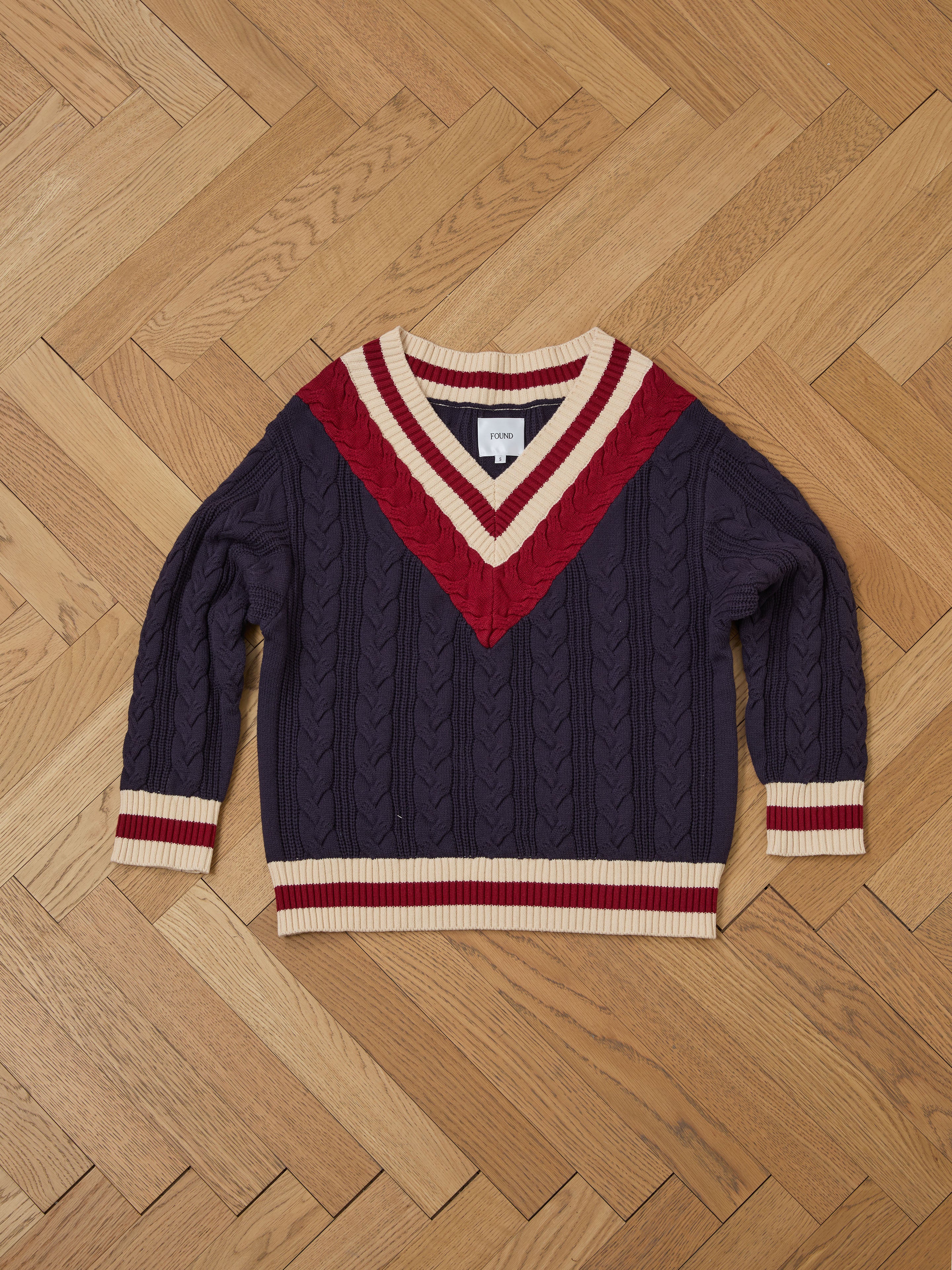 The FOUND V-Neck Cricket Sweater features a cable-knit design with red and cream accents at the neck and cuffs, elegantly showcased on a wooden parquet floor.