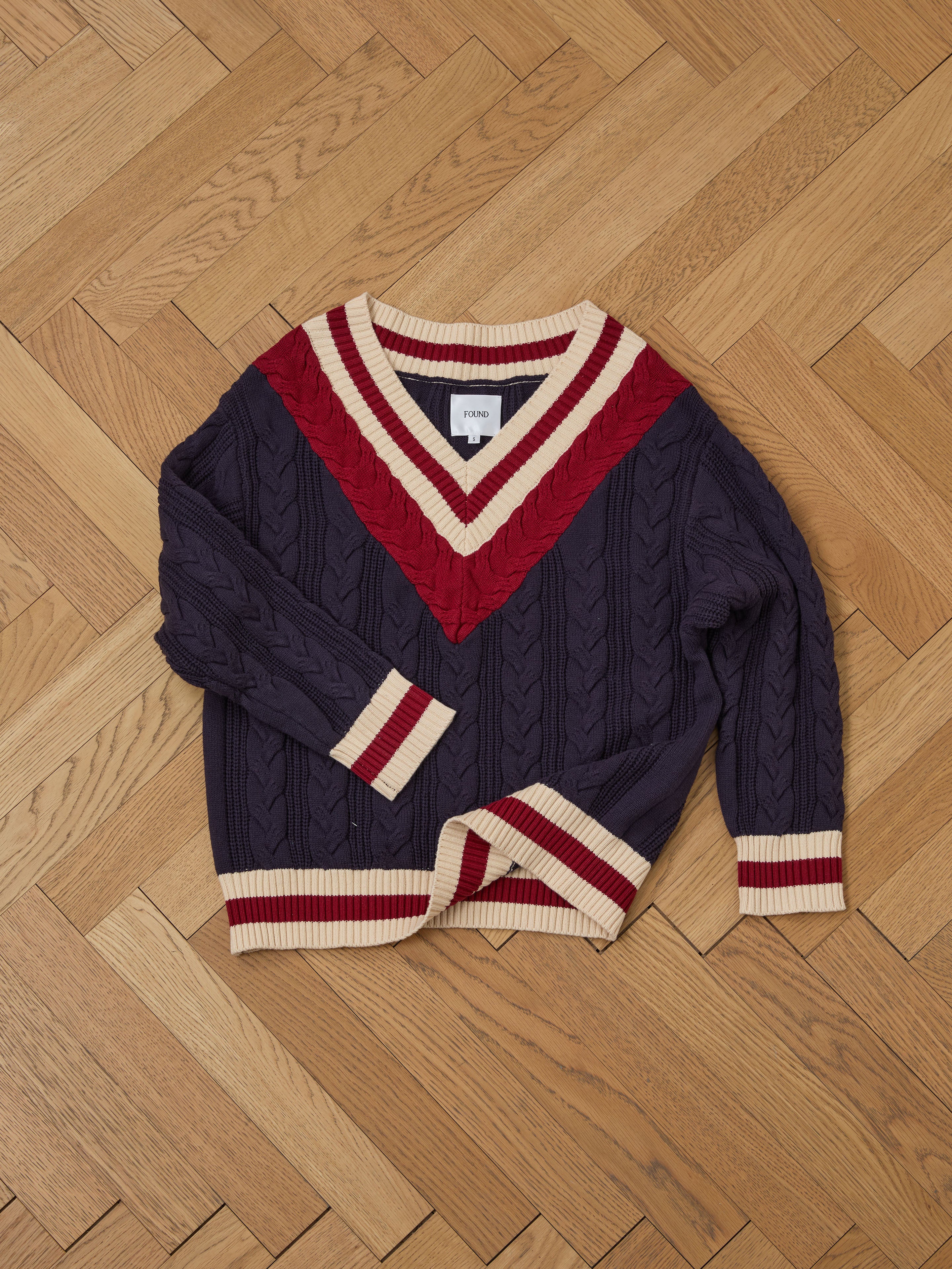 A navy FOUND V-Neck Cricket Sweater with red and cream stripes, crafted from soft cotton, sits on a wooden herringbone-patterned floor.