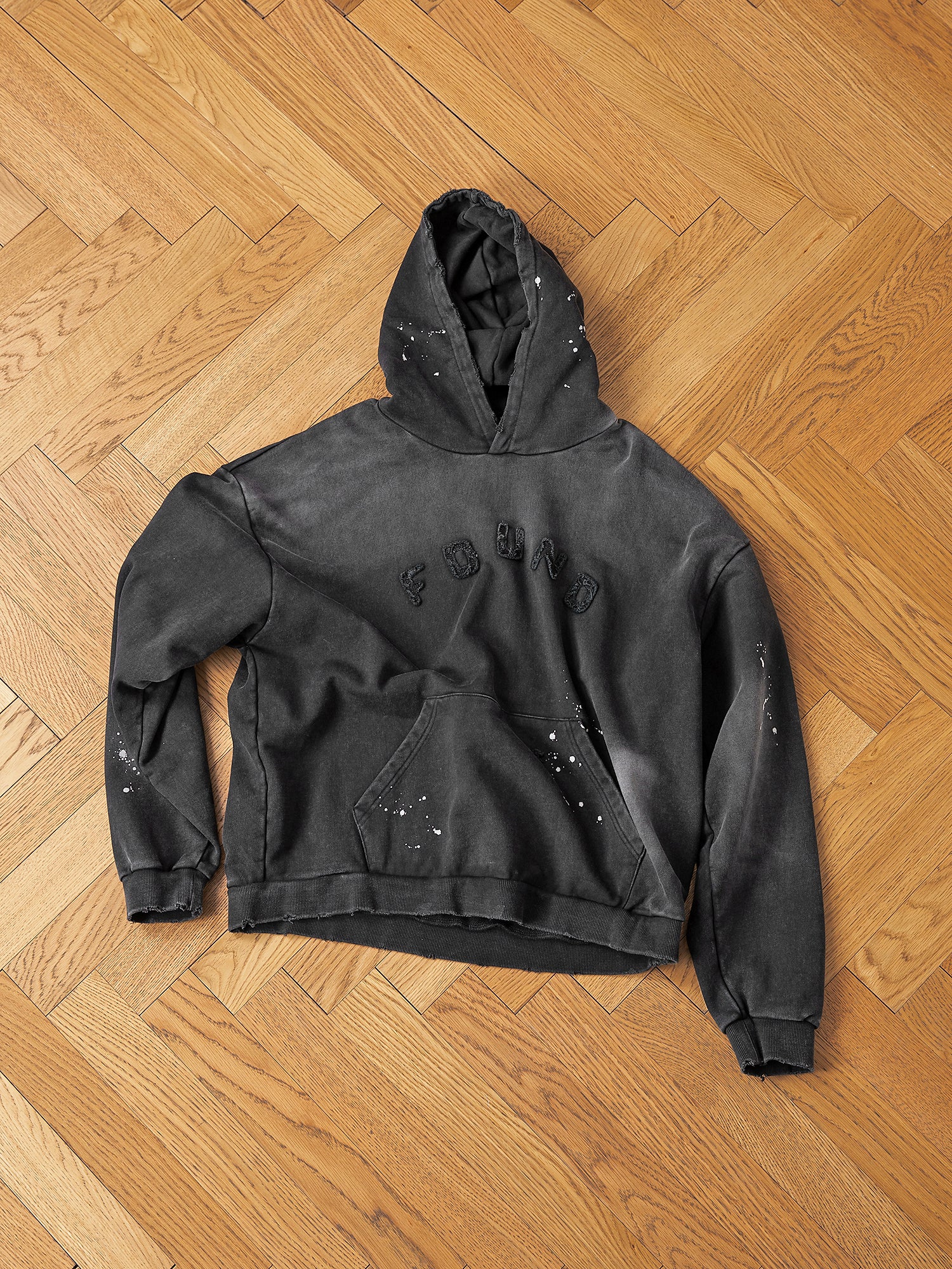 A Logo Patch Hoodie from FOUND, crafted in vintage black from soft French terry cotton, displays "FOUND" across the chest. It features a unique distressed design with artistic paint splatters, beautifully set on a wooden parquet floor.