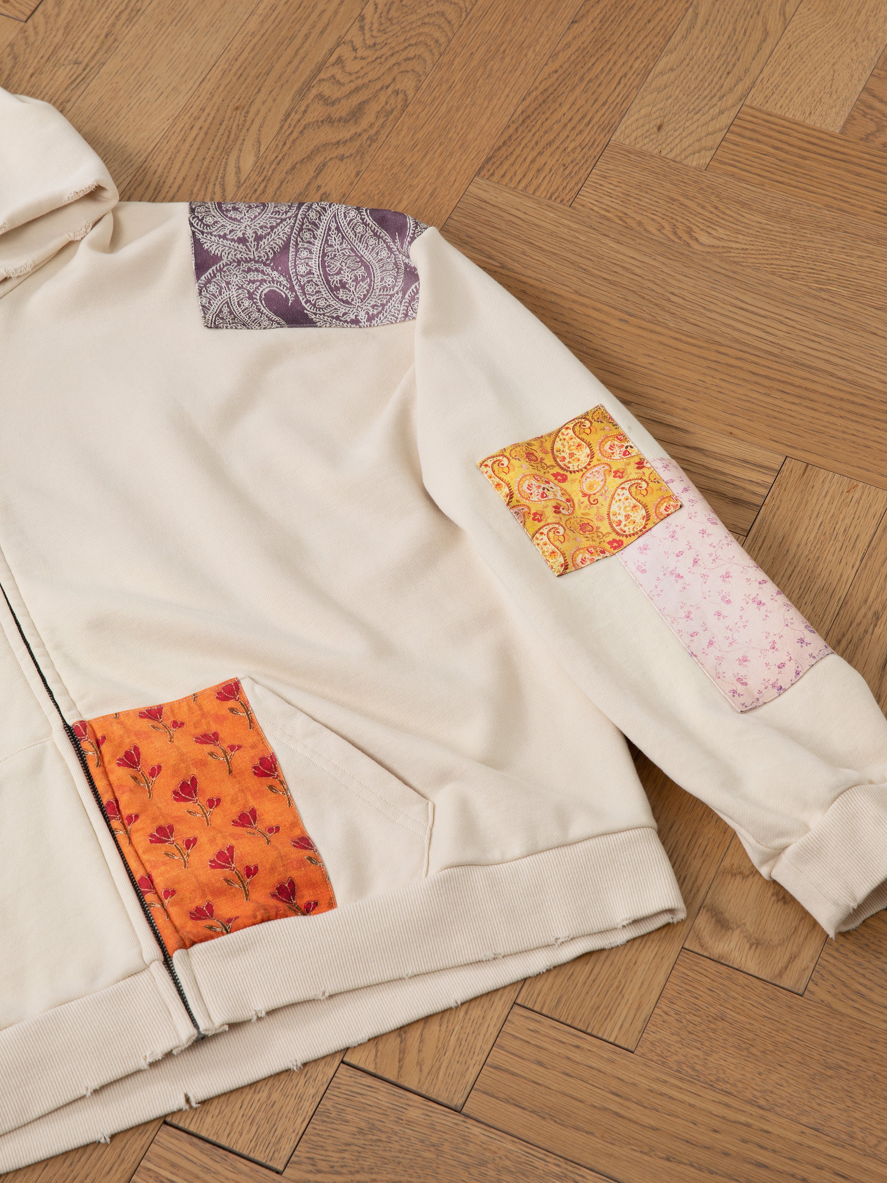 The Tapestry Patchwork Zip Up Hoodie from FOUND features a cream design crafted with French terry cotton and vibrant South Asian tapestry patterns in purple, yellow, orange, and pink adorning the chest, sleeve, and side. It is elegantly displayed on a wooden floor.