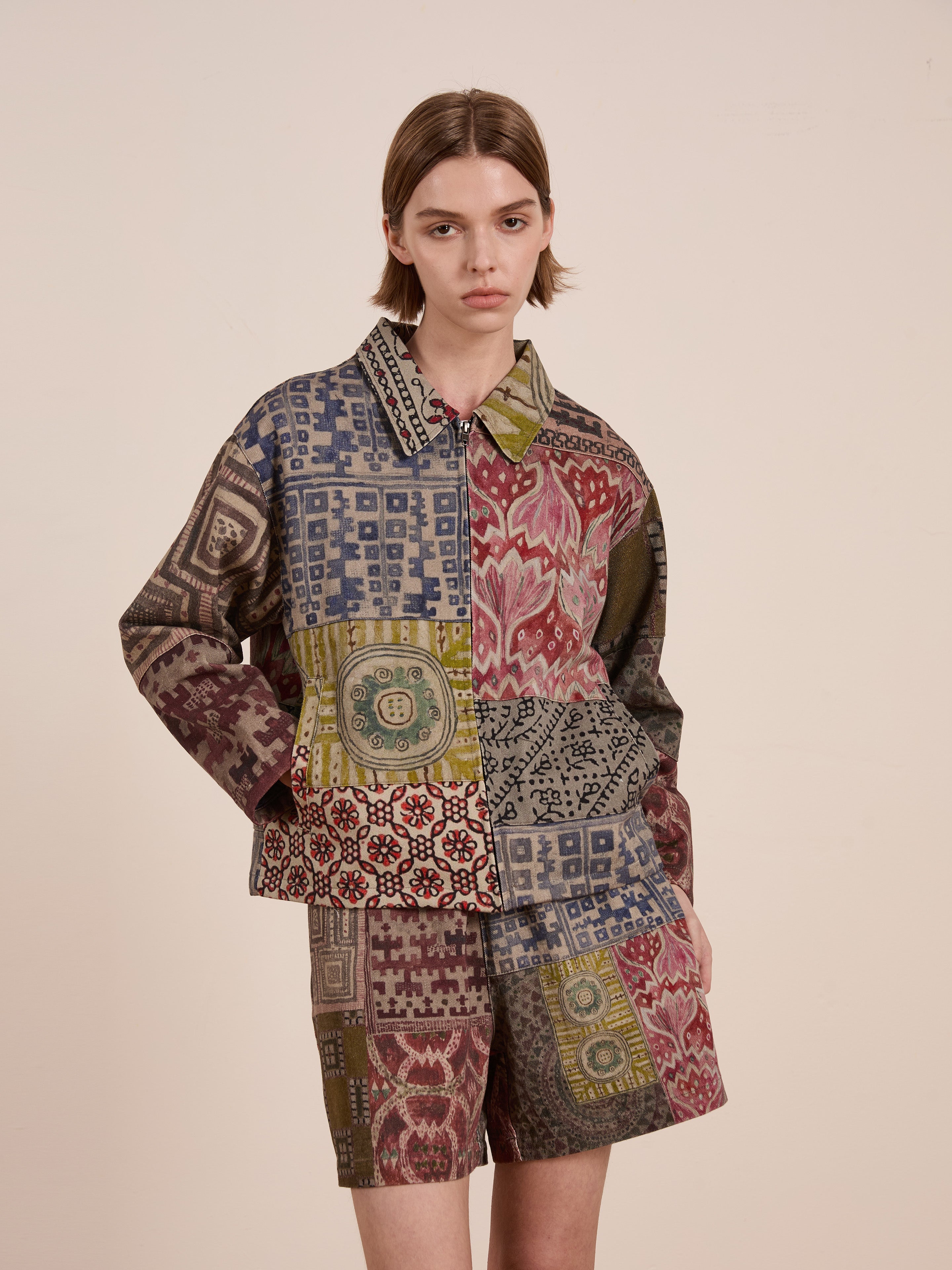 Against a plain beige background, a person wears FOUND's vibrant Watercolor Mosaic Work Jacket and shorts. The unisex, long-sleeved design bursts with creativity and color, showcasing true artistic flair.