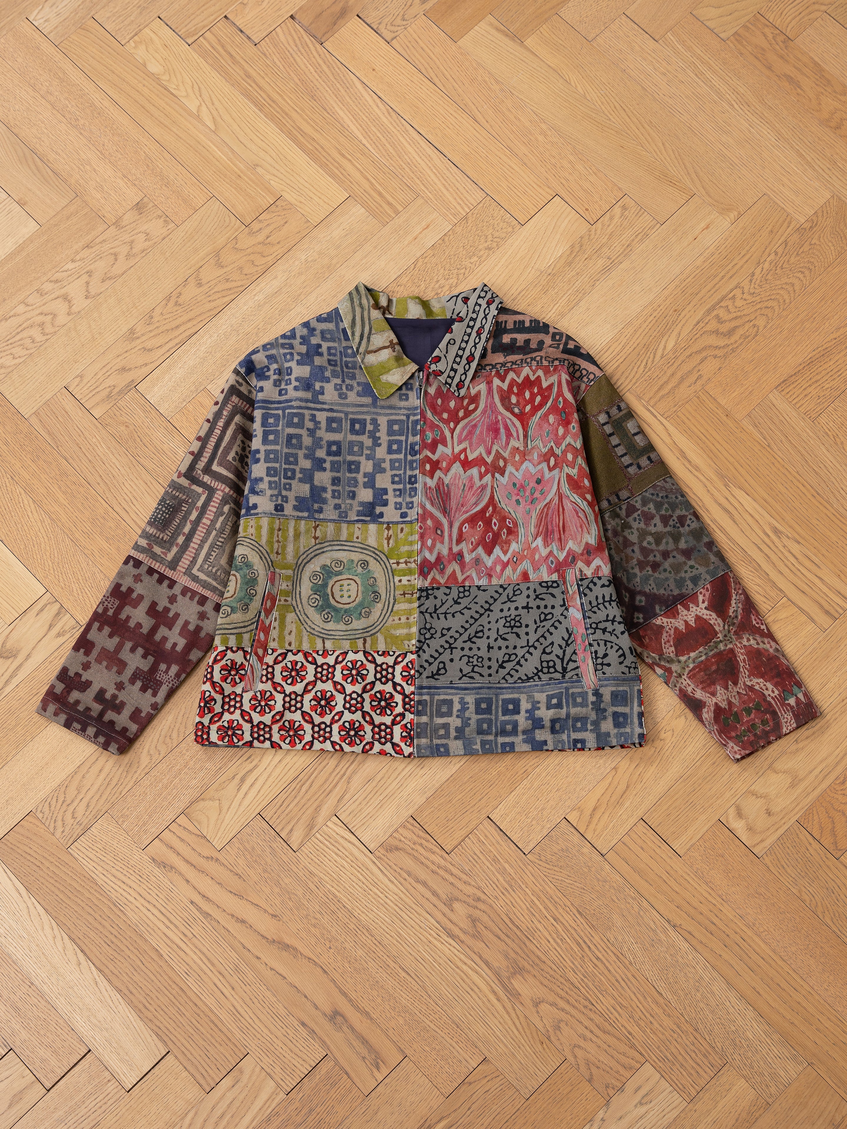 The Watercolor Mosaic Work Jacket by FOUND is a unisex, long-sleeve jacket showcasing vibrant patchwork with geometric and floral patterns, expertly styled on wooden parquet. It effortlessly combines style and versatility for any wardrobe.