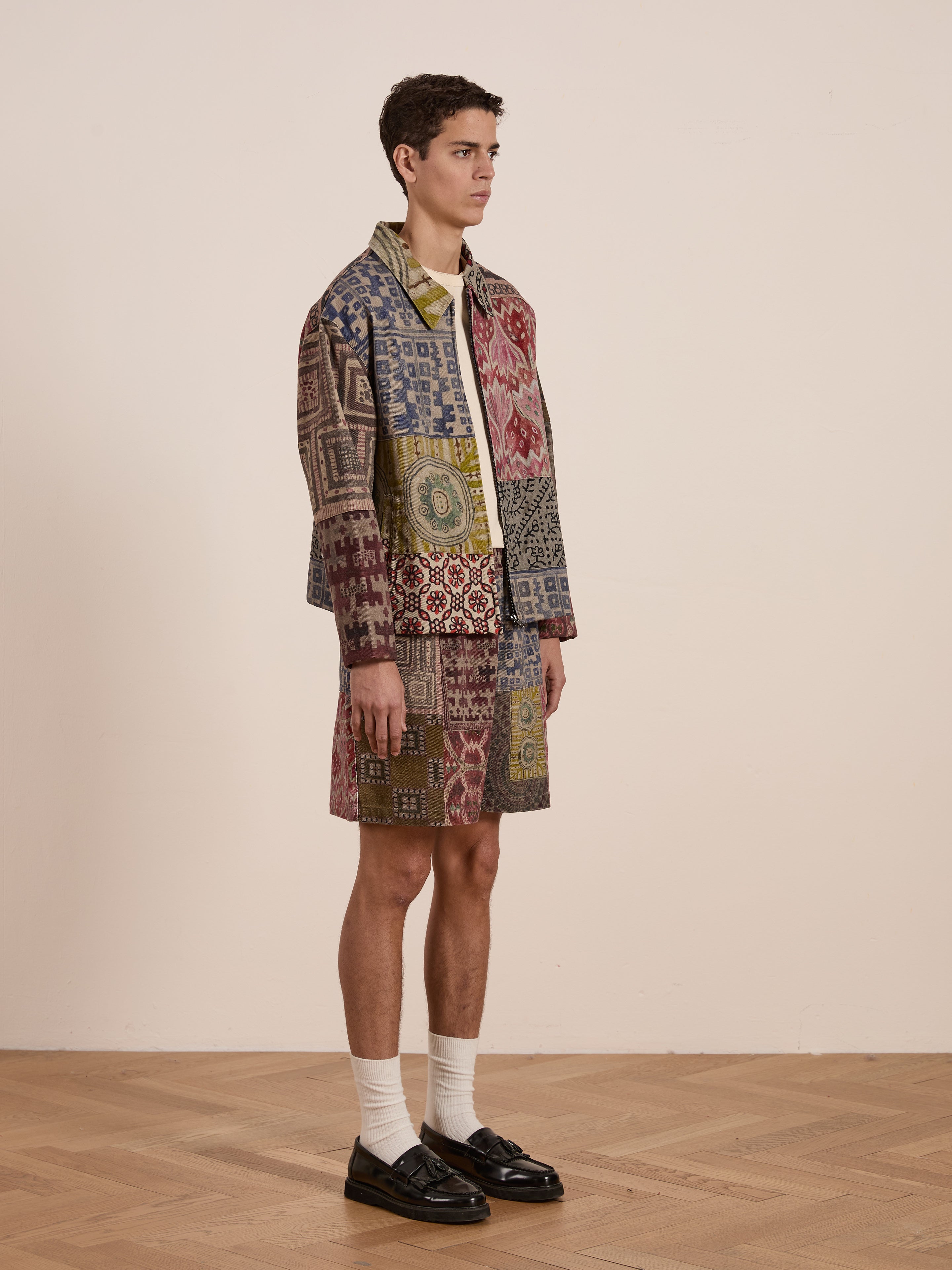 In a room, someone wears the FOUND Watercolor Mosaic Work Jacket and matching shorts featuring intricate watercolor patterns. White socks peek from their black shoes as they gaze sideways, highlighting the bold style of this unisex ensemble.