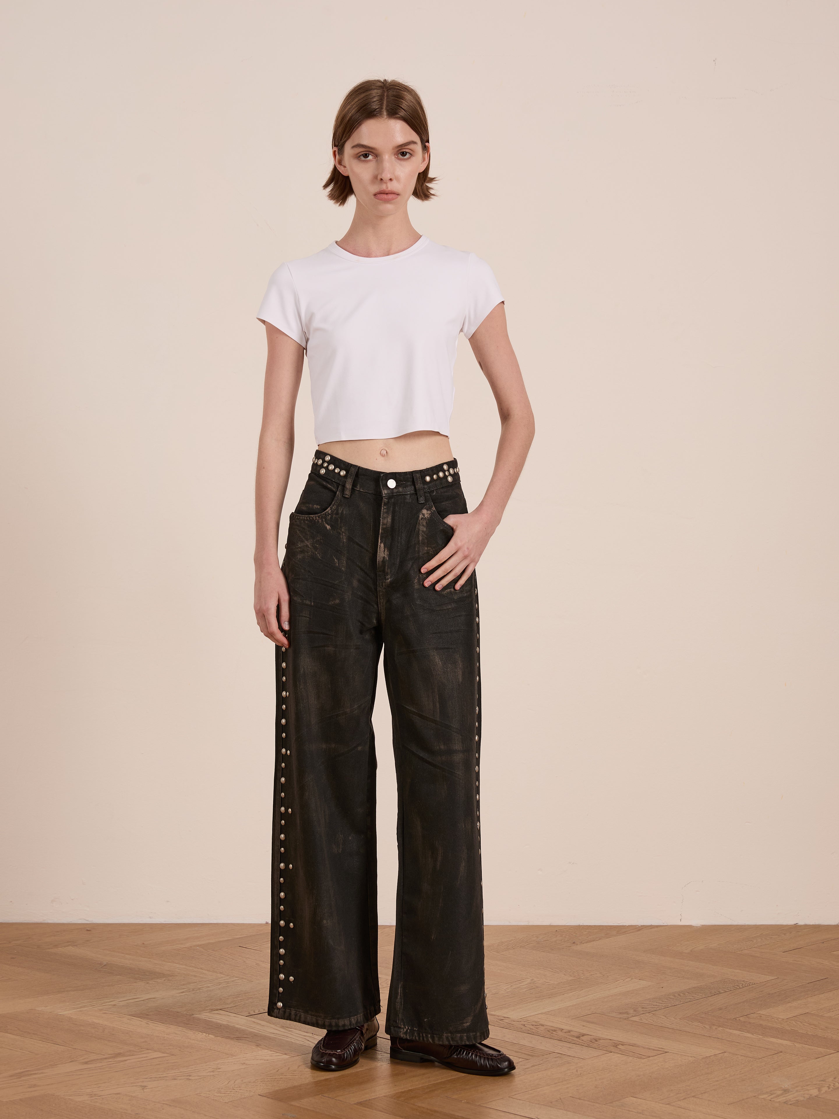 A person wearing a white crop top and FOUND's Waxed Studded Jeans stands on a wooden floor against a plain beige wall.