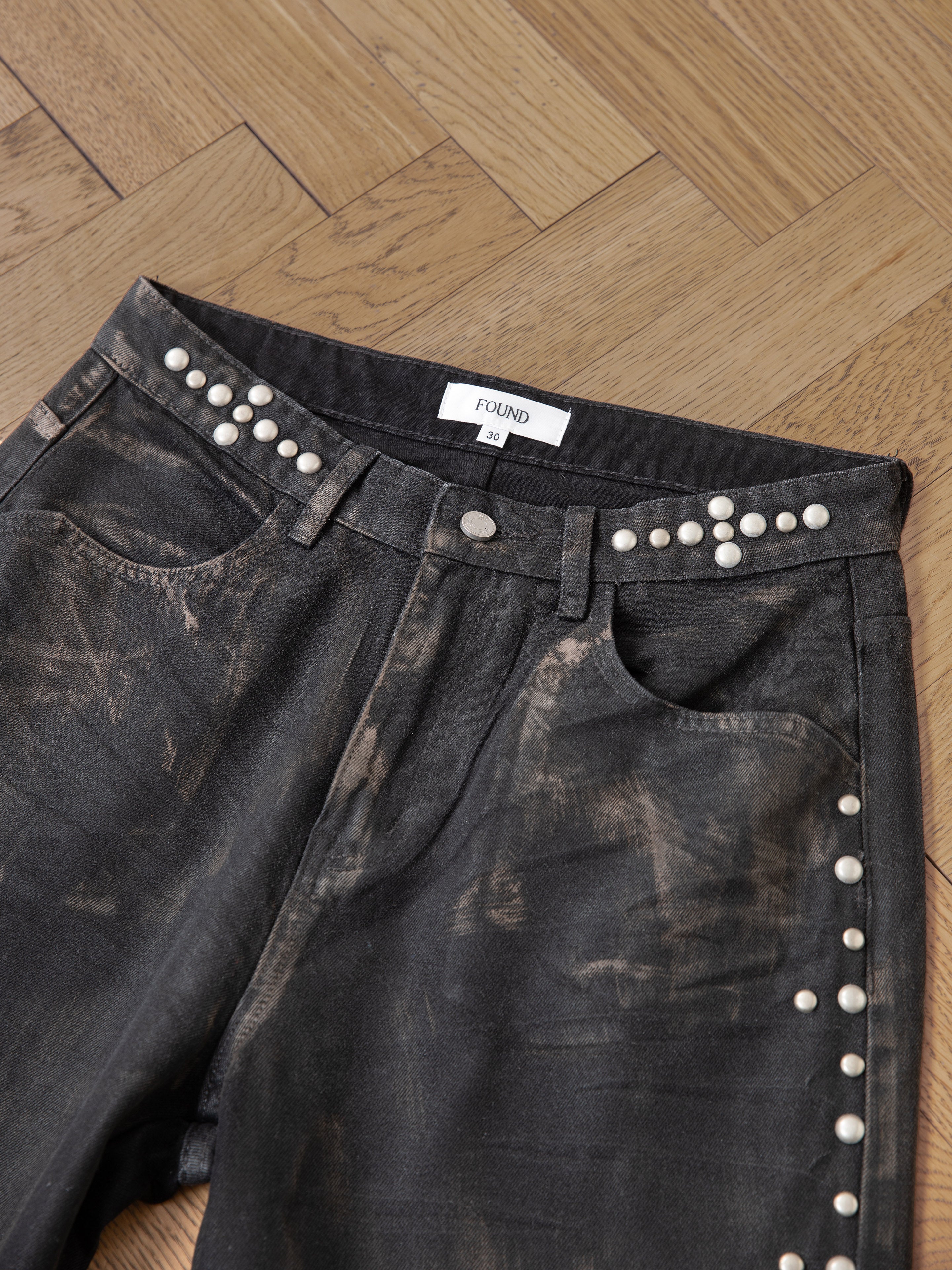 Waxed Studded Jeans by FOUND: Black denim featuring vintage craftsmanship with a distressed look, studded waist and pockets. Unisex relaxed fit showcased on a wooden floor.