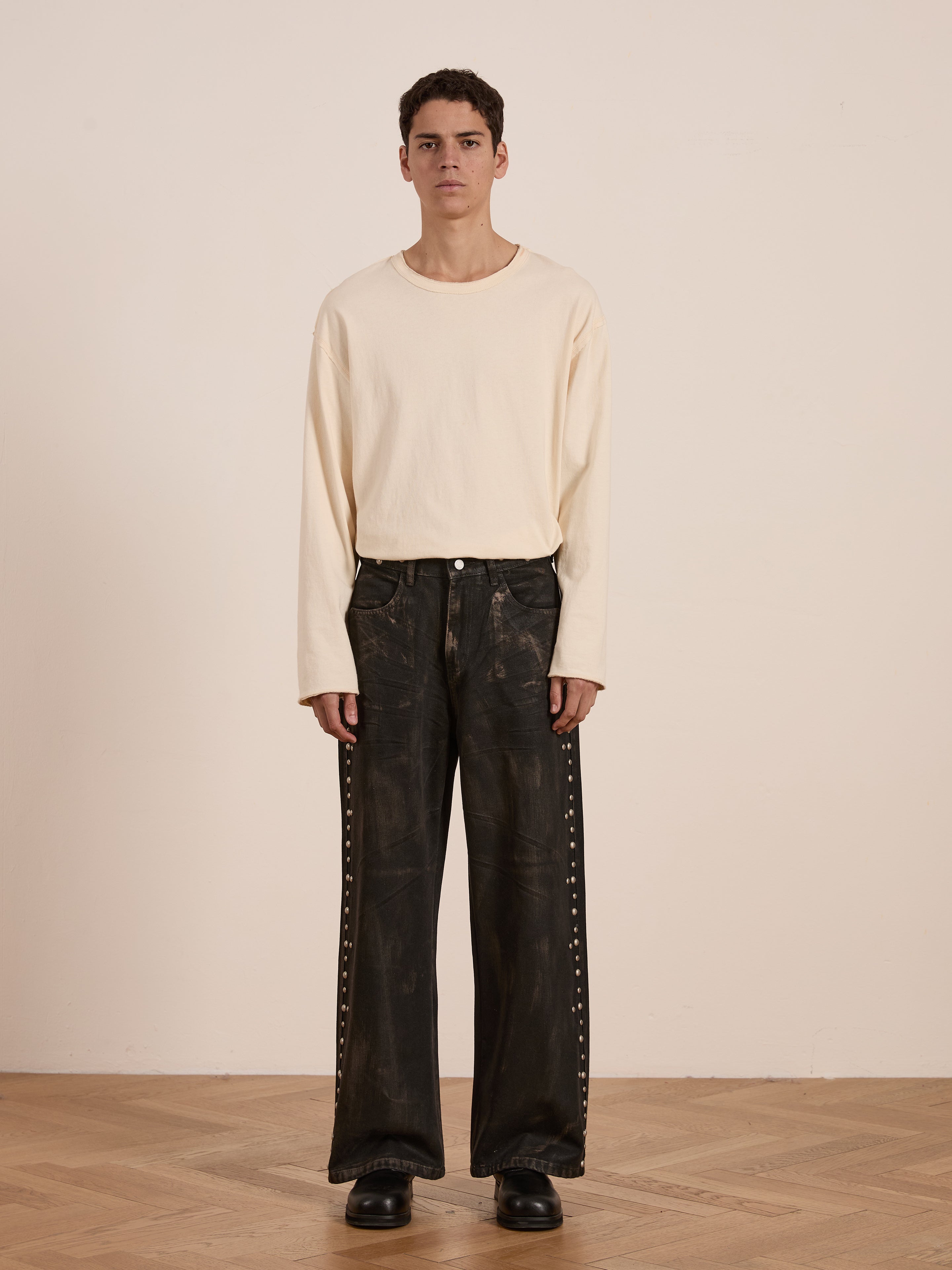 Someone stands on a wooden floor, wearing a cream long-sleeve shirt and FOUND’s Waxed Studded Jeans, unisex relaxed-fit dark pants with vintage craftsmanship against a plain background.