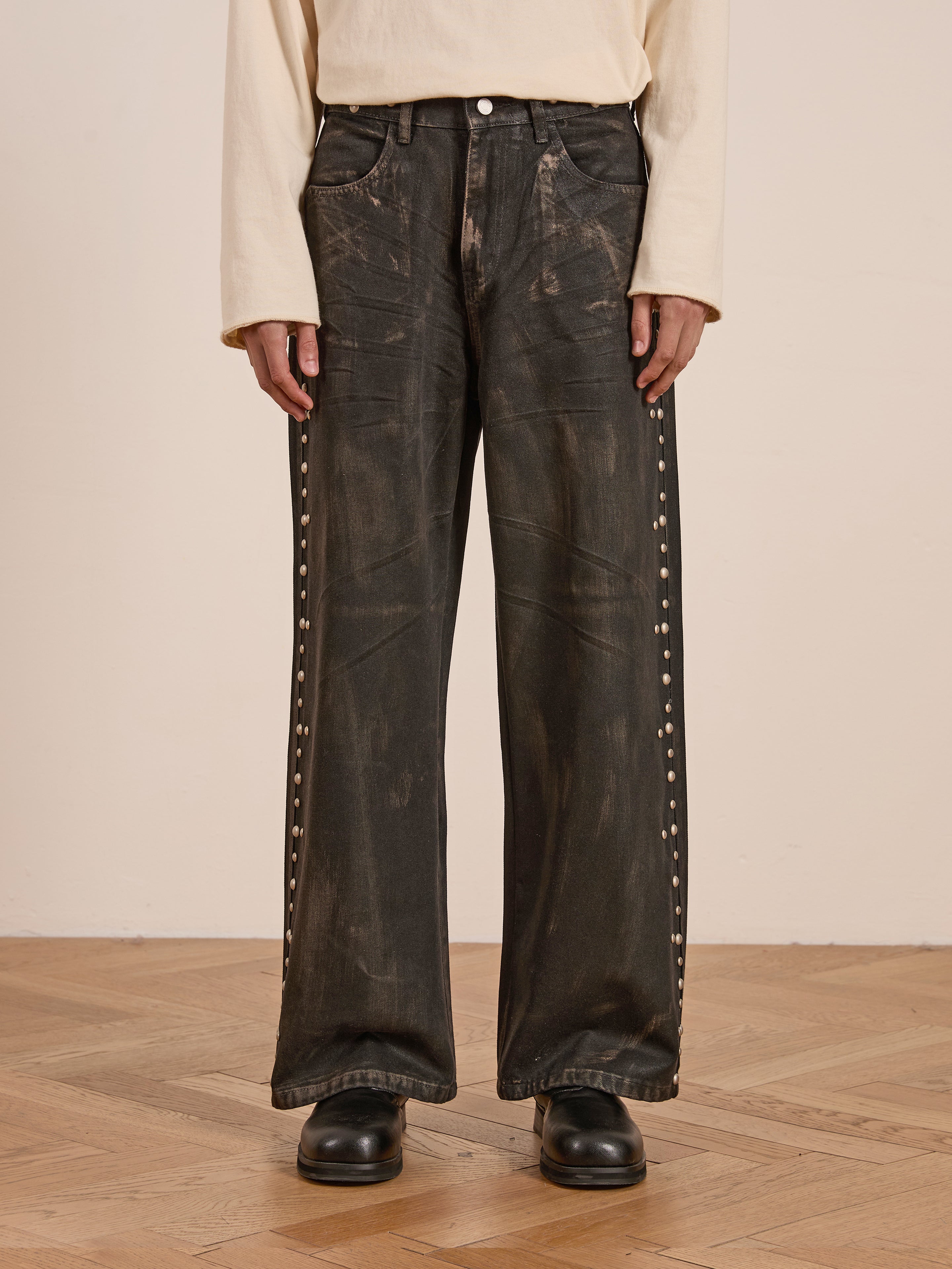 A person stands on a wooden floor, wearing Waxed Studded Jeans from FOUND that exude vintage craftsmanship, paired with a beige long-sleeve shirt.