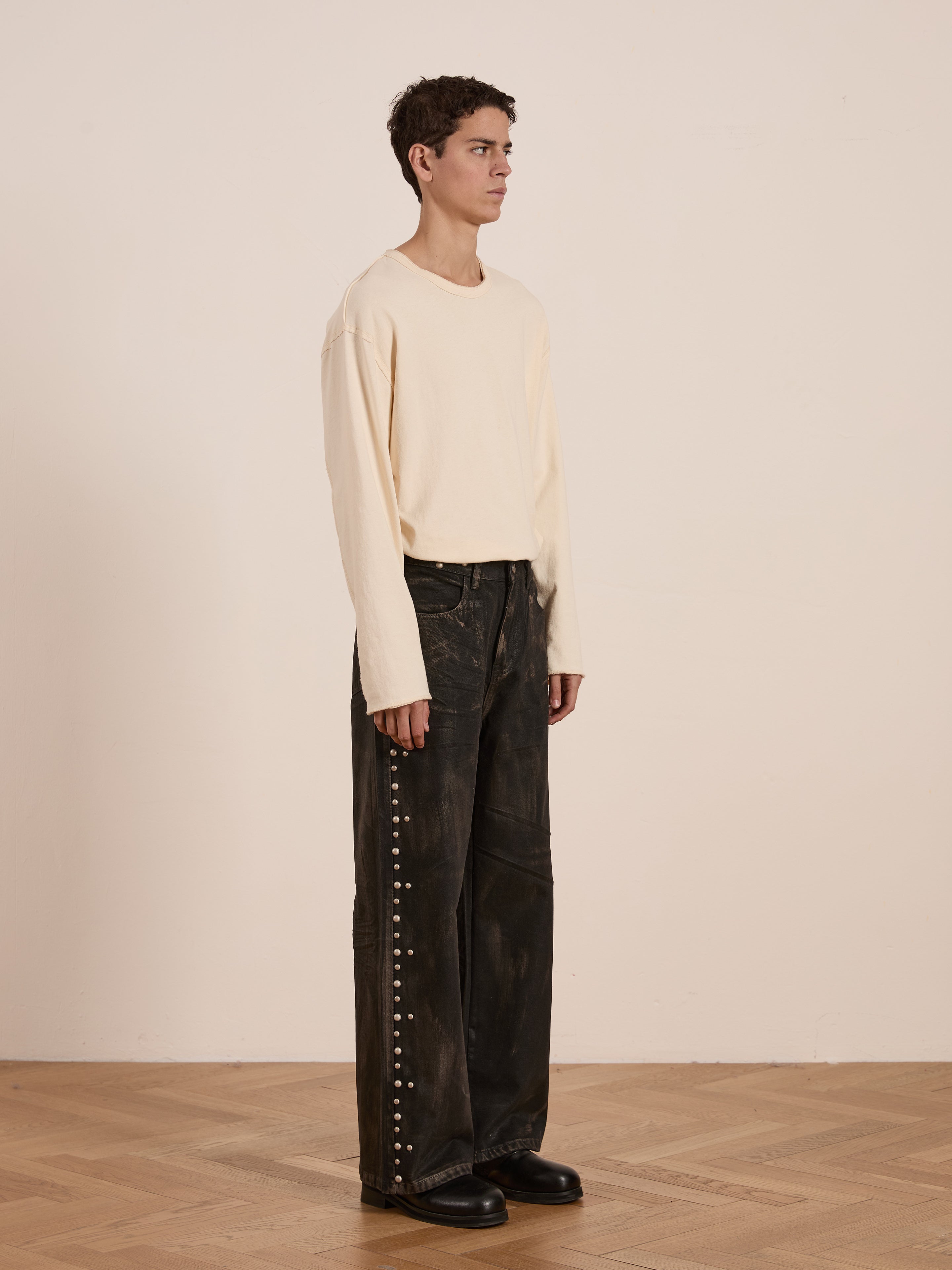 A person stands on a wooden floor wearing FOUND's Waxed Studded Jeans, paired with a long-sleeve beige shirt and sleek black shoes. Their vintage-inspired outfit stands out against an off-white wall.