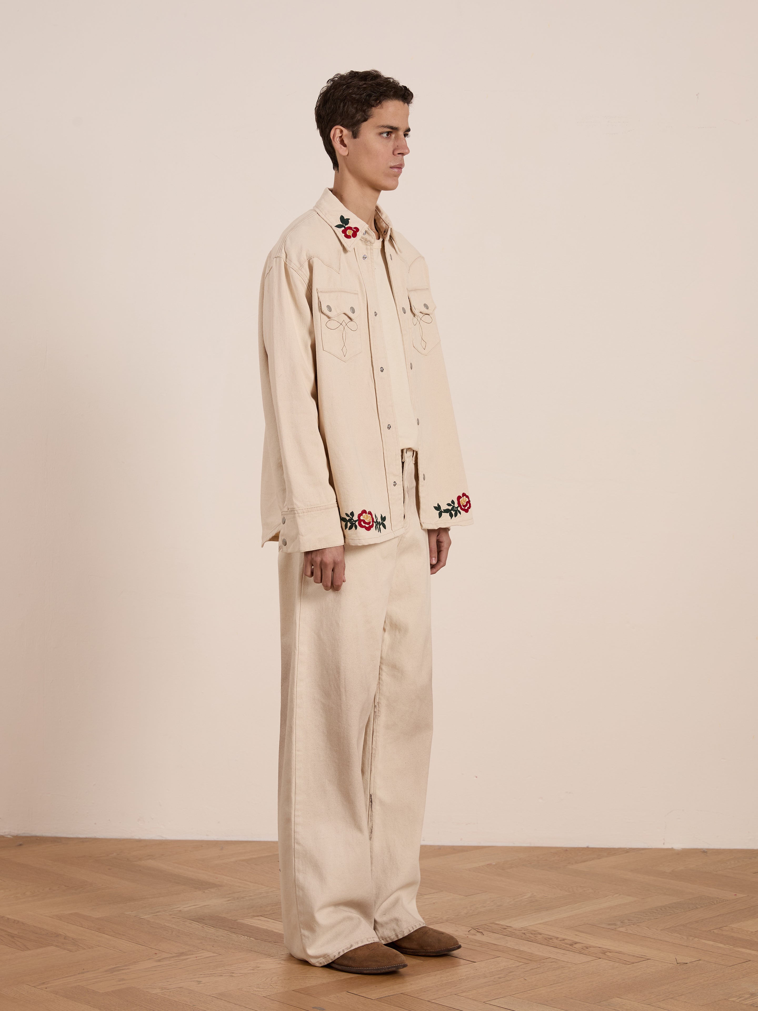 A person in profile on a wooden floor showcases the FOUND Western Floral Denim Shirt, featuring beige fabric with floral chainstitch embroidery against a plain backdrop.