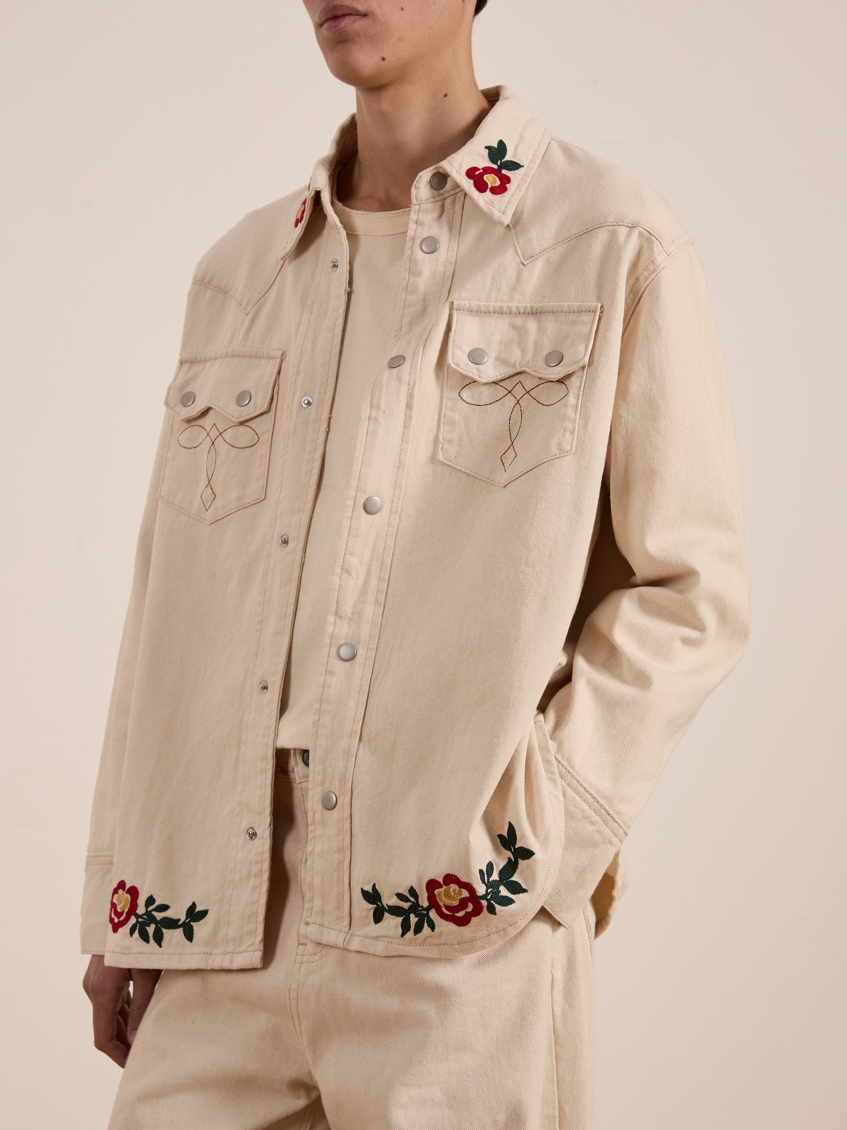 A person is wearing FOUND's Western Floral Denim Shirt in beige, featuring floral chainstitch embroidery on the collar and pockets, paired with matching pants.