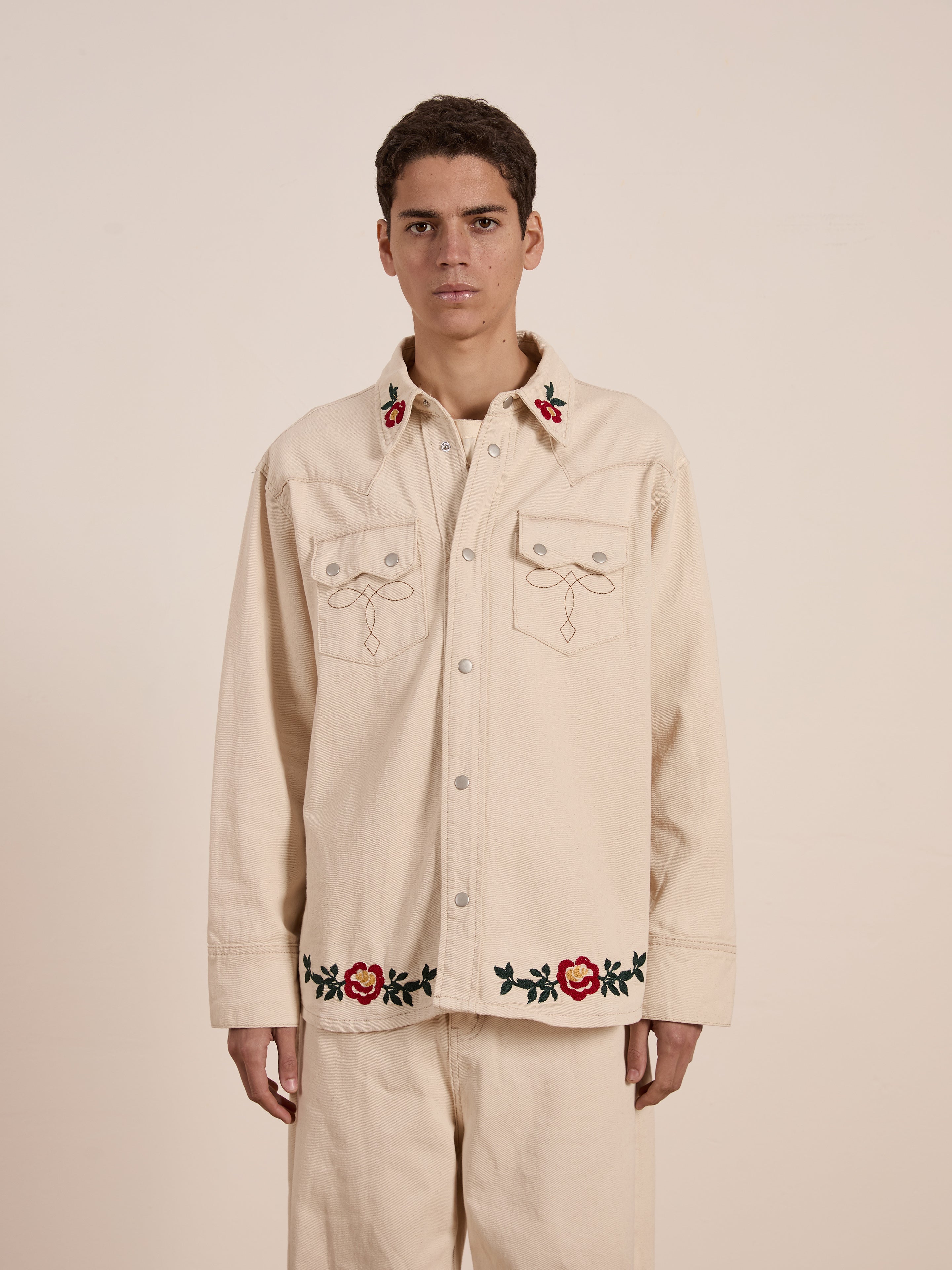 Against a plain backdrop, someone wears a Western Floral Denim Shirt by FOUND, in cream with detailed floral chainstitch embroidery on the collar and hem.