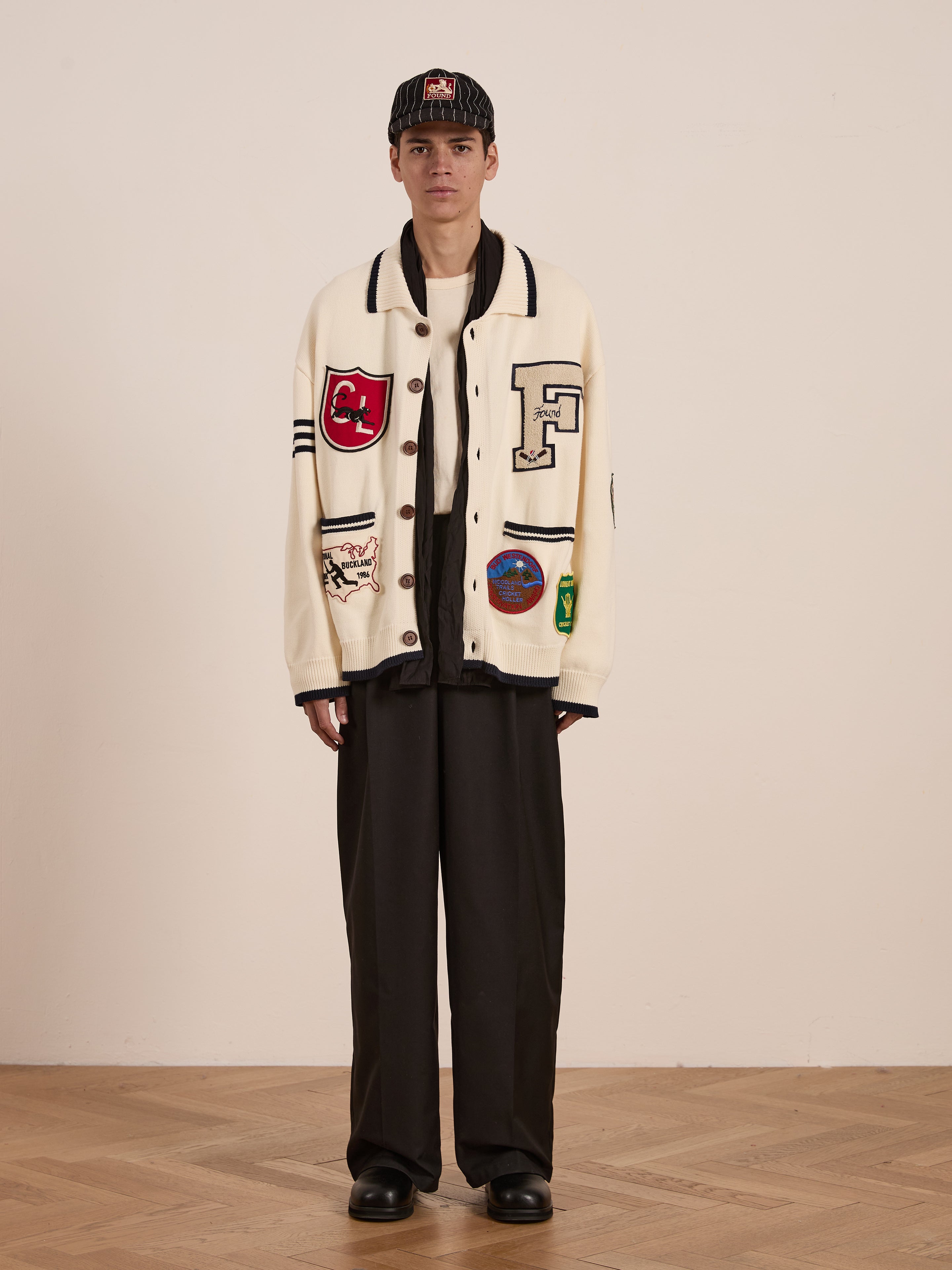 A person in a FOUND York Varsity Patch Collared Cardigan, black wide-leg pants, and a black cap stands on a wooden floor against a plain backdrop, exuding preppy style with collegiate nostalgia.