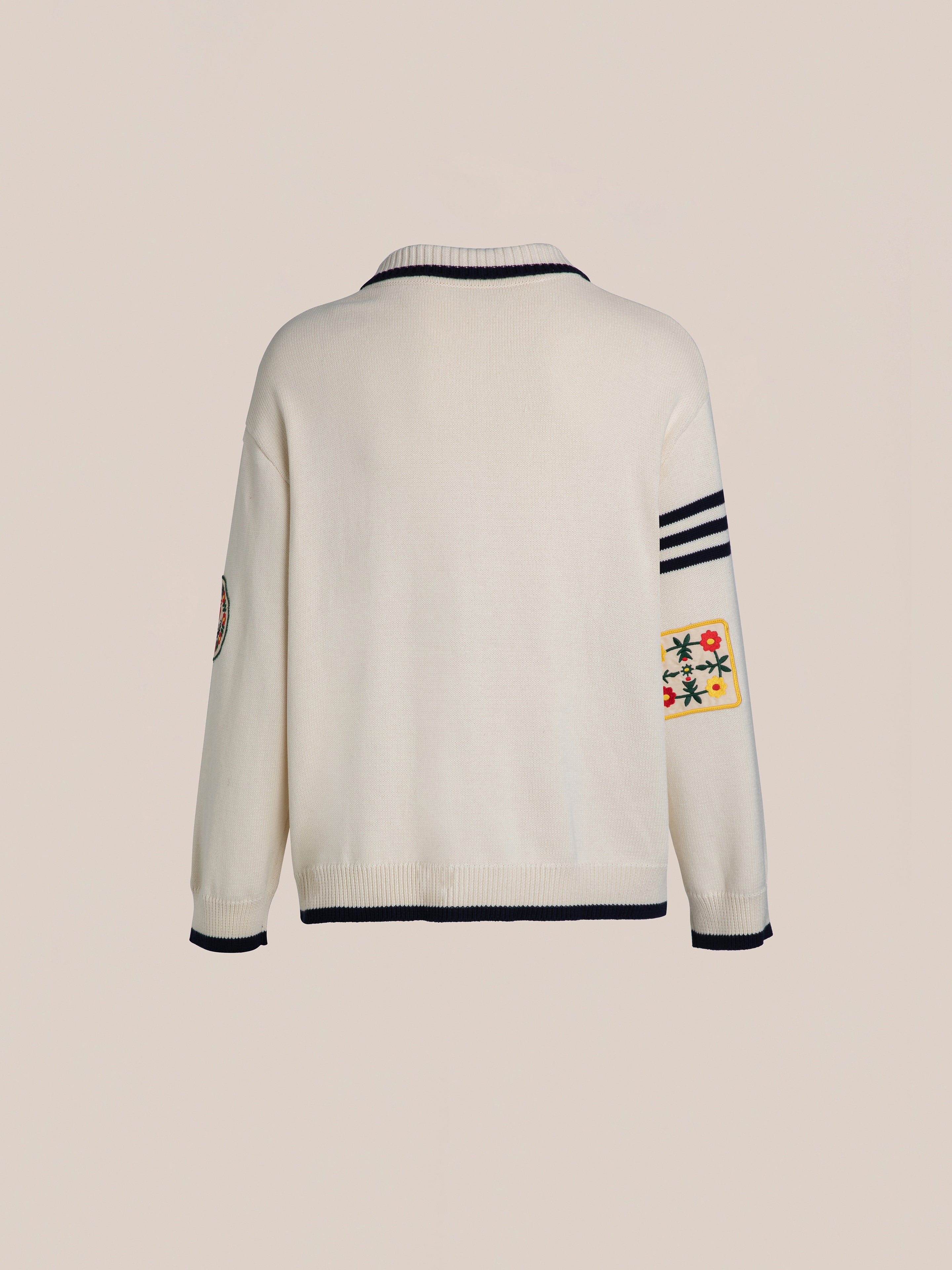 The York Varsity Patch Collared Cardigan by FOUND is a white sweater with black trim, boasting multicolored floral embroidery on the right sleeve and three varsity-style black stripes on the left, capturing a preppy collegiate aesthetic against a plain backdrop.