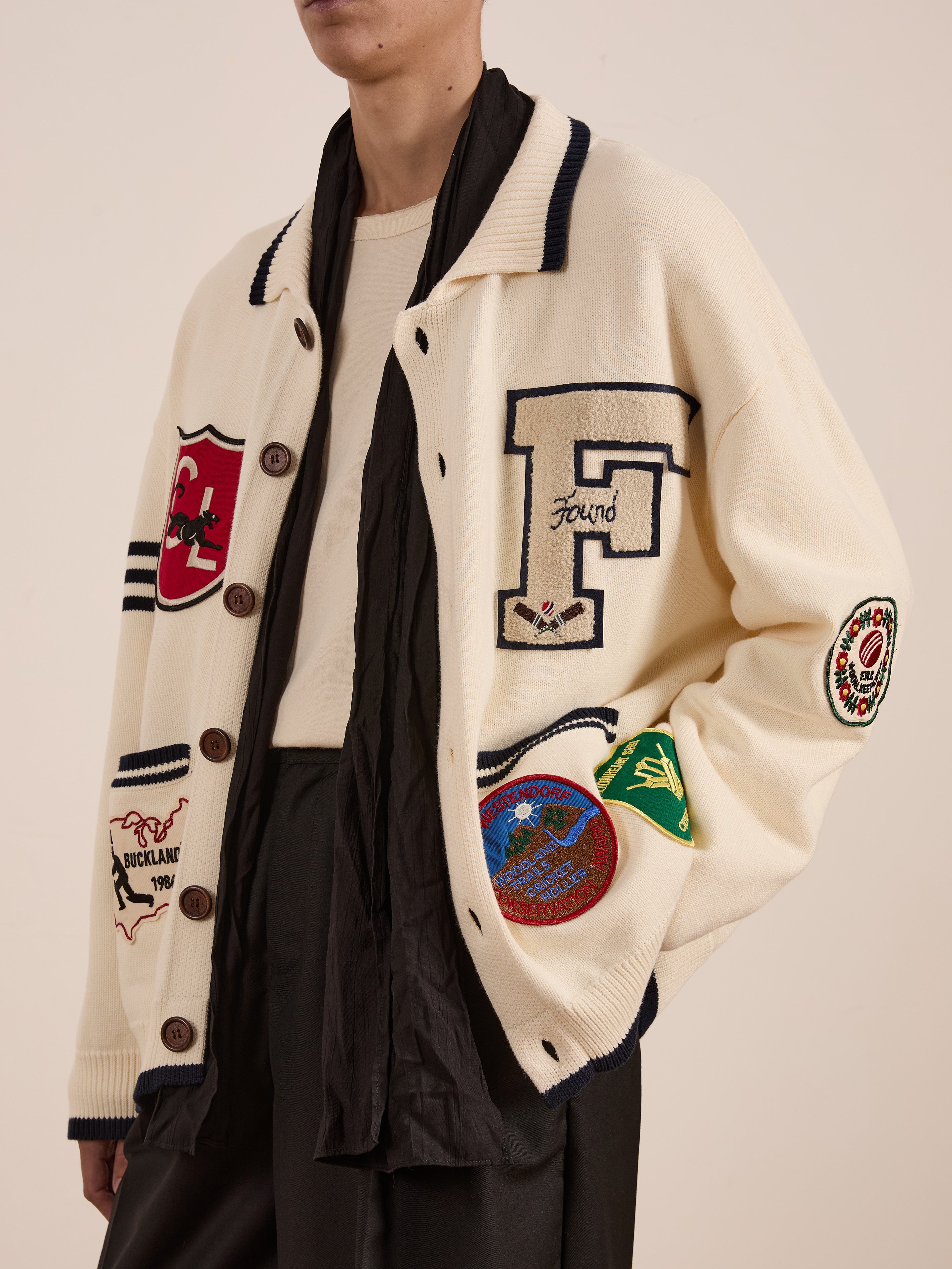 A person wearing a York Varsity Patch Collared Cardigan by FOUND, featuring a prominent "F" on the front, embodies the preppy style. The look is enhanced with a black scarf and dark pants, evoking collegiate nostalgia.