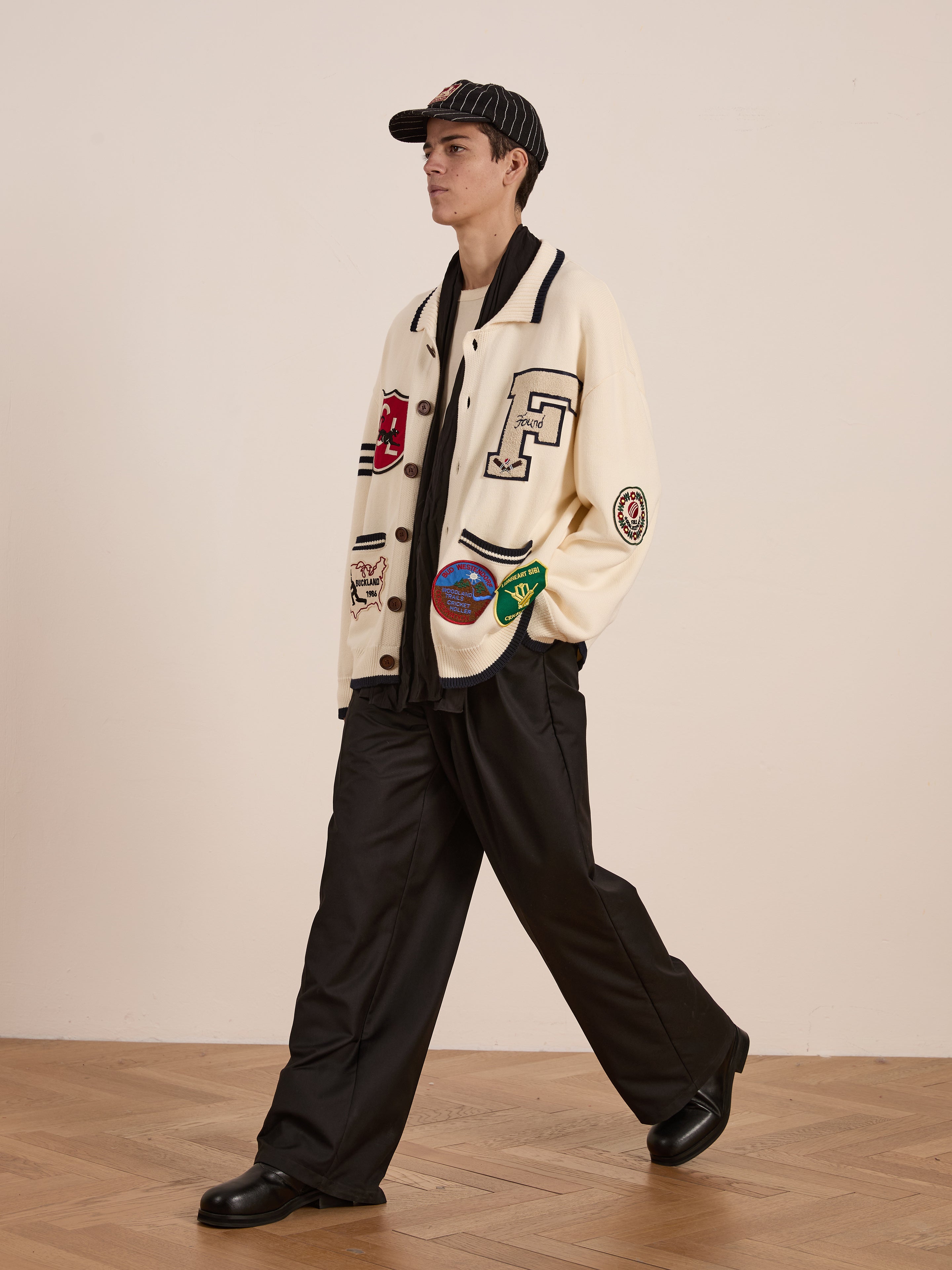 With collegiate nostalgia, a person walks indoors on a wooden floor, wearing FOUND's York Varsity Patch Collared Cardigan, black wide-leg pants, a black shirt, and a striped cap.