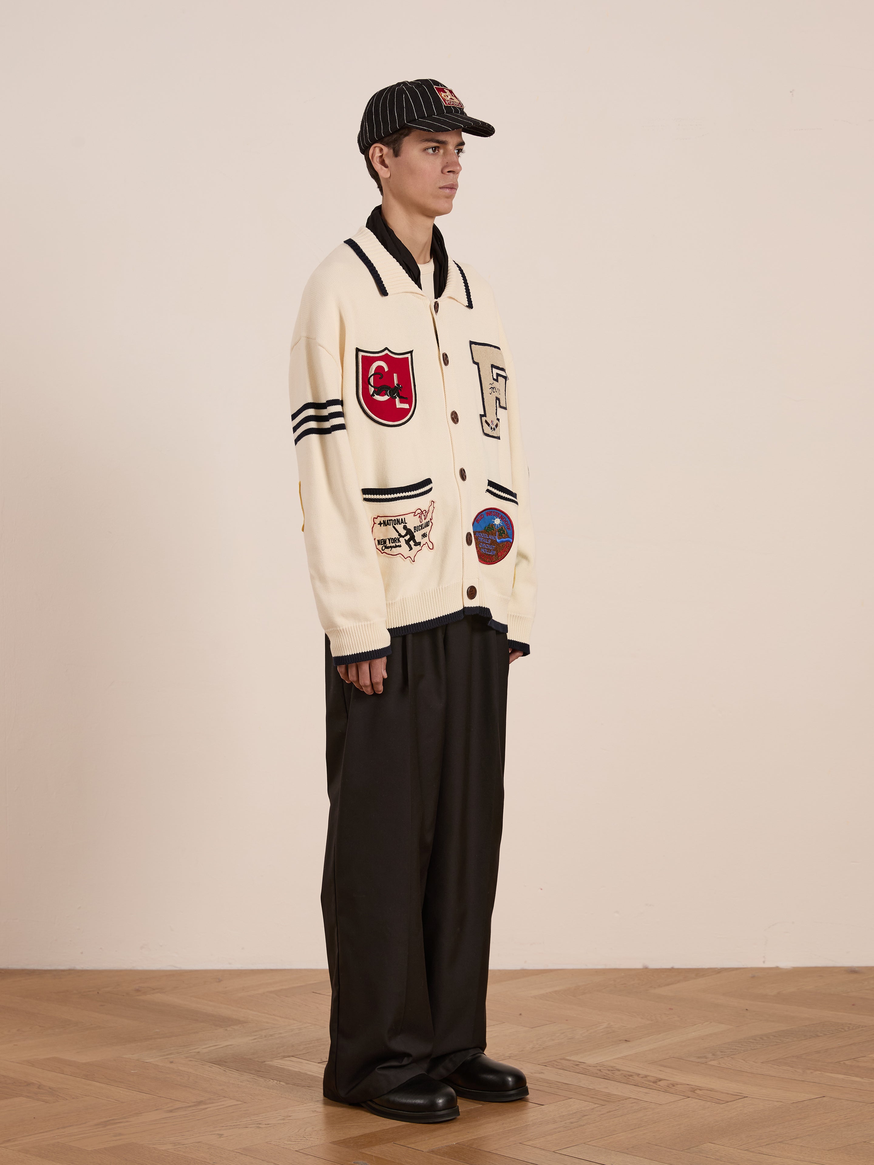 A person in profile exudes collegiate nostalgia in a preppy York Varsity Patch Collared Cardigan by FOUND, paired with black wide-leg trousers. The ensemble is completed with a striped cap against the backdrop of a neutral-toned room.