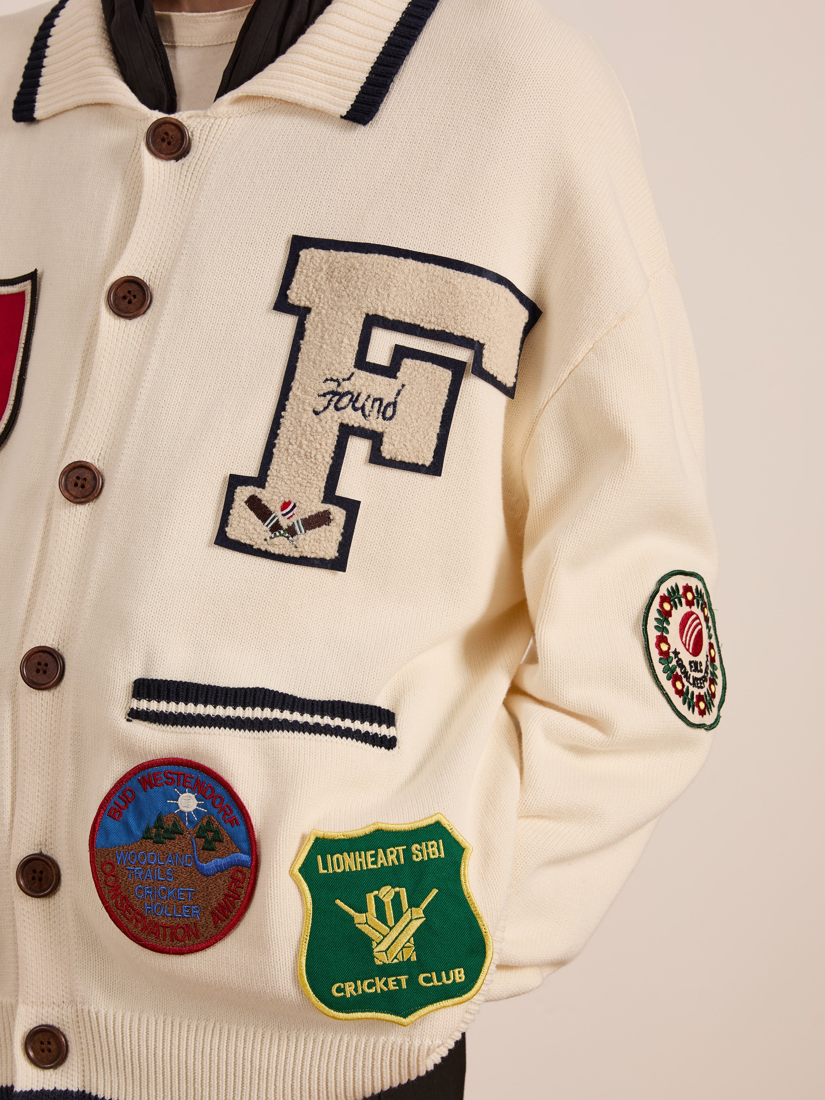 A person is wearing FOUND's York Varsity Patch Collared Cardigan, a cream-colored jacket featuring a large "F" and colorful cricket-themed patches, offering a preppy vibe with collegiate nostalgia.