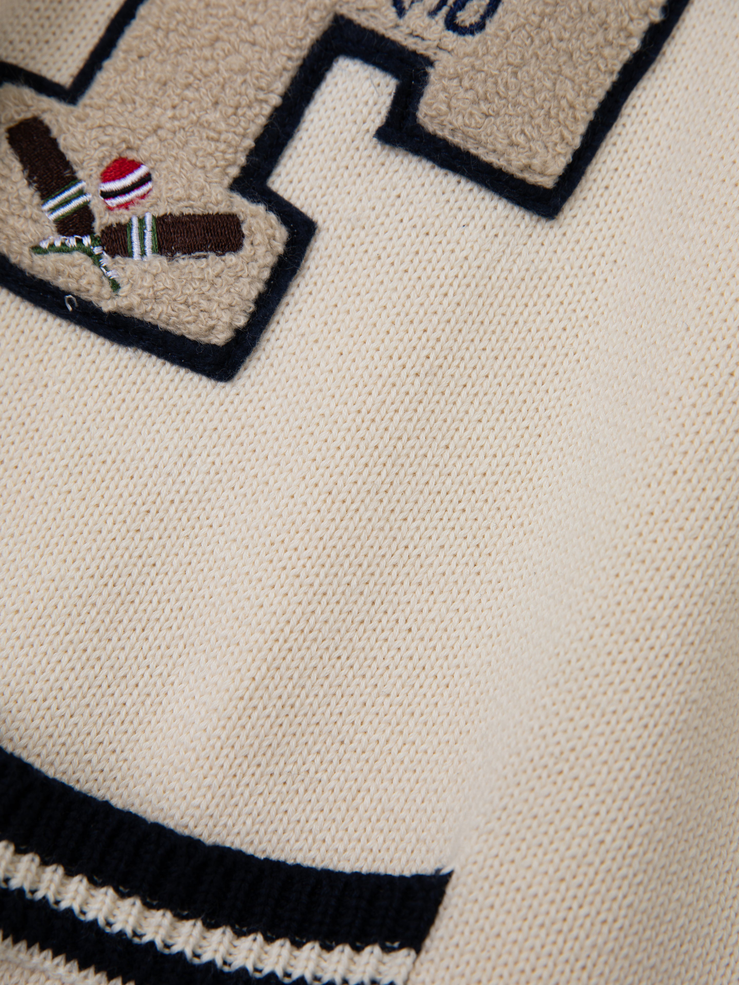 The York Varsity Patch Collared Cardigan by FOUND features a close-up beige textured fabric with a stitched 'H', crossed cricket bats, ball design, and navy stripes. It's perfect for a preppy, collegiate aesthetic reminiscent of varsity style.