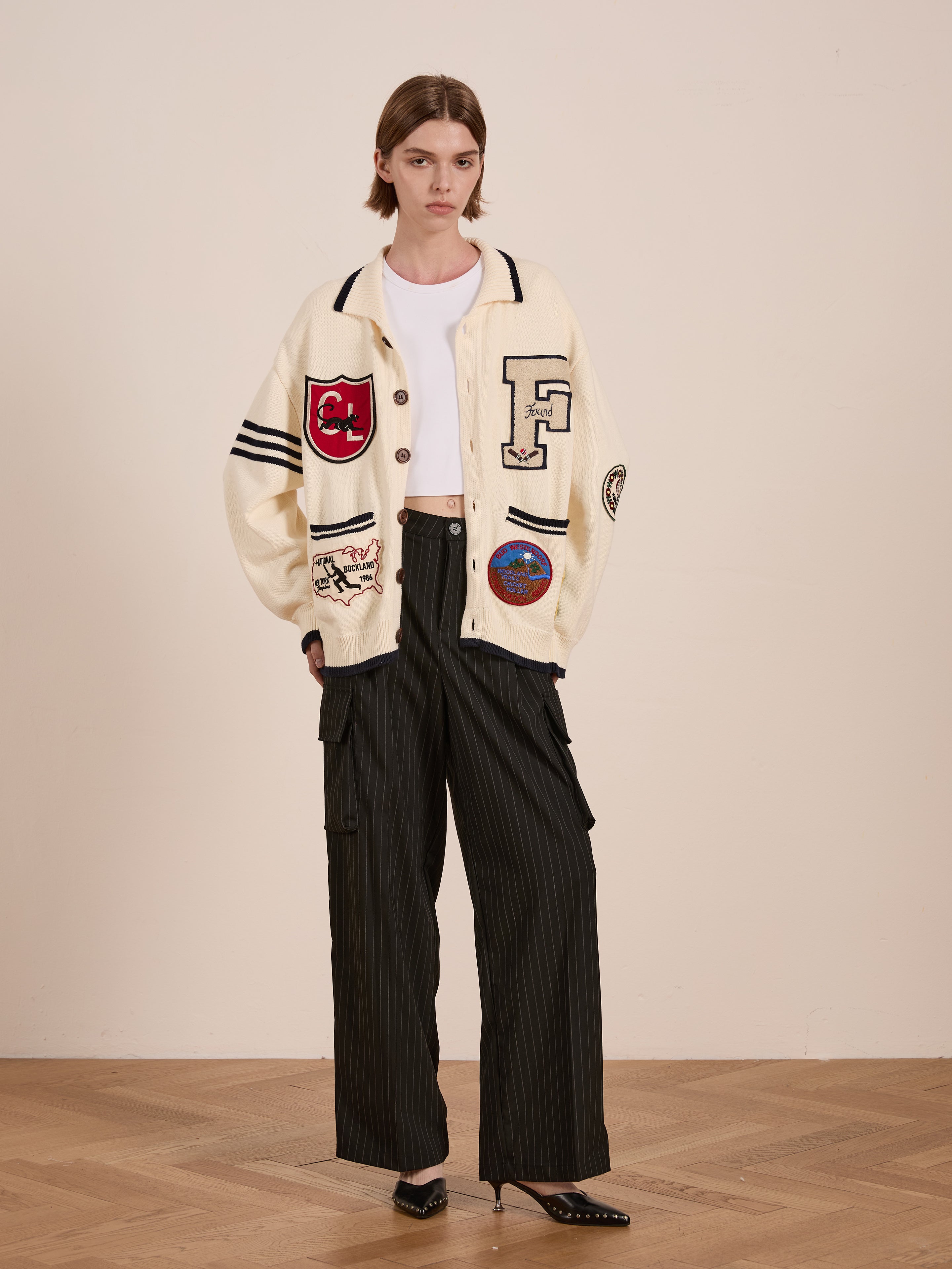 A person stands on a wooden floor wearing FOUND's York Varsity Patch Collared Cardigan, exuding collegiate nostalgia, paired with a white crop top and black pinstripe cargo pants.